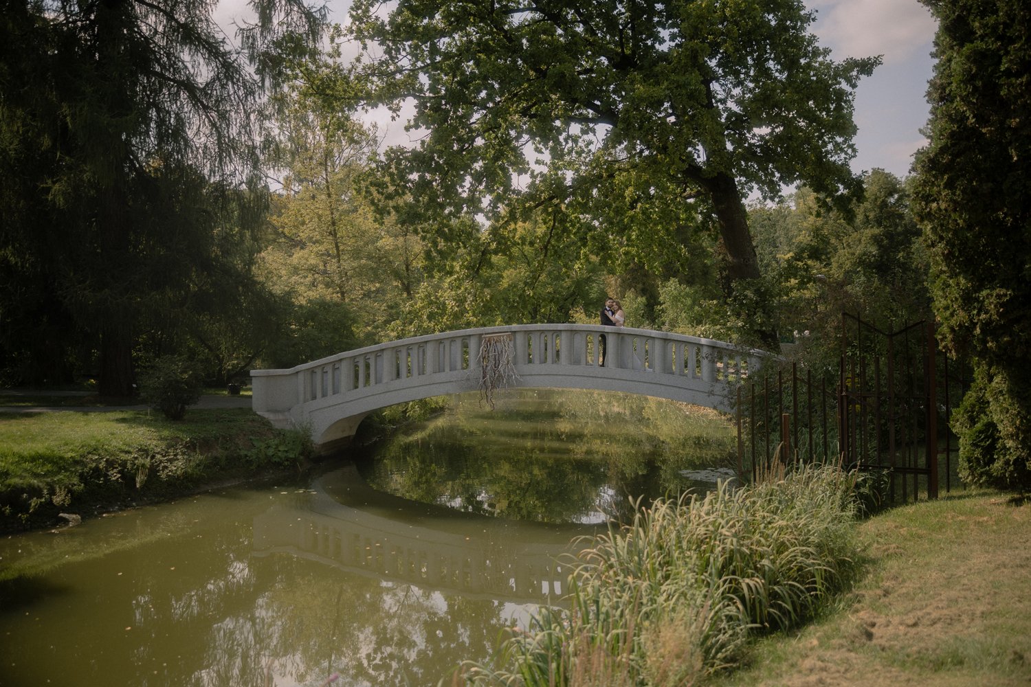 A white curved footbridge over a small river in a park, surrounded by green trees and foliage, with two people standing on the bridge.