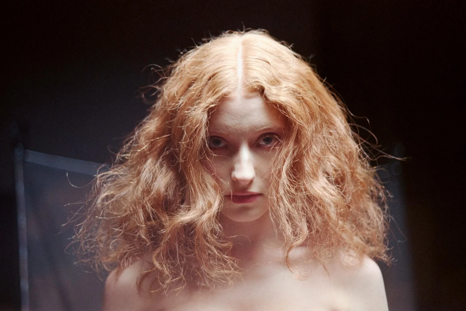 A woman with curly red hair looking directly at the camera against a dark background.