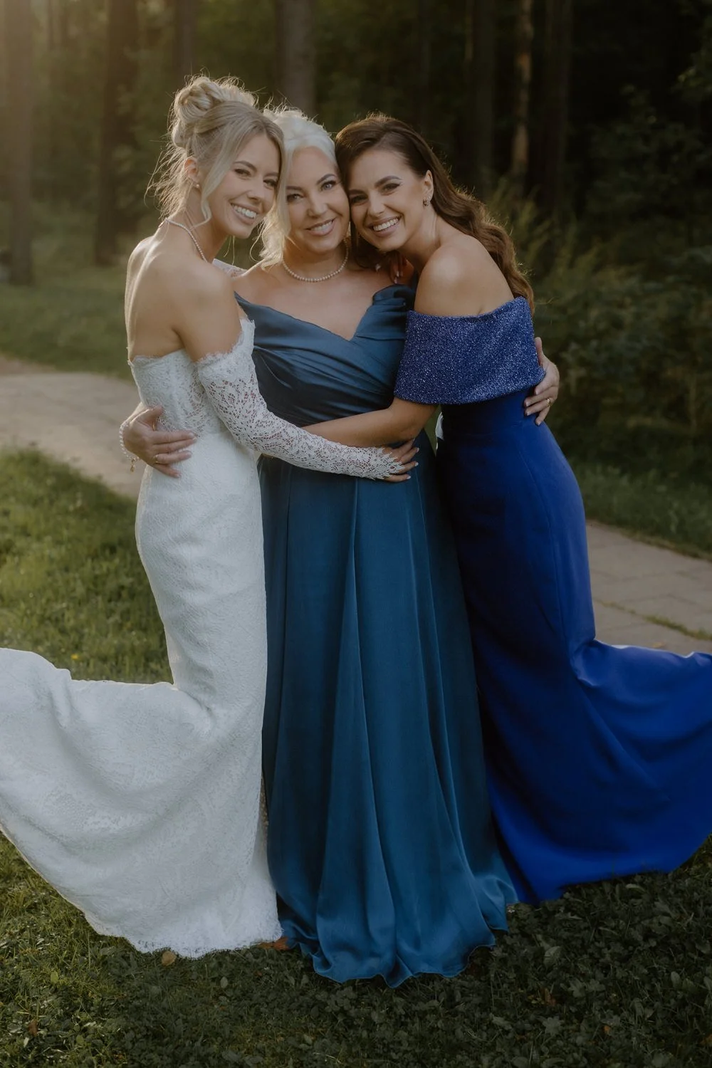Three women dressed in elegant gowns, smiling and embracing outdoors in a forested area with greenery and sunlight