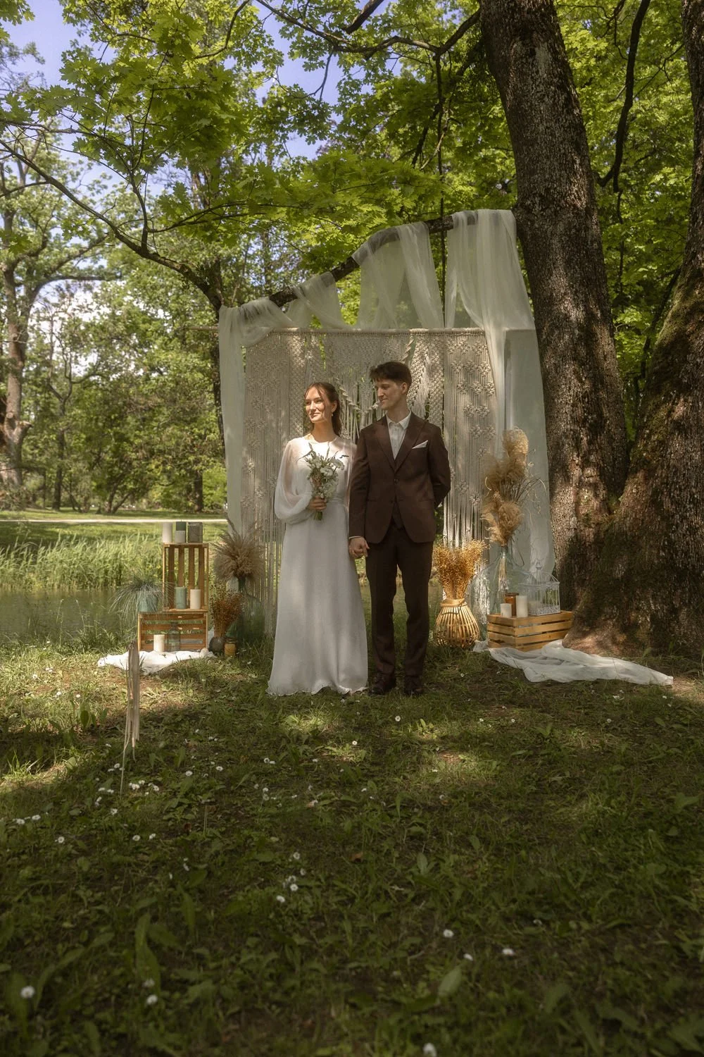 A bride and groom standing together outdoors in a wooded area during a wedding ceremony. The bride is holding a bouquet and wearing a white dress, while the groom is in a dark suit. They are under a canopy decorated with sheer fabric and surrounded b