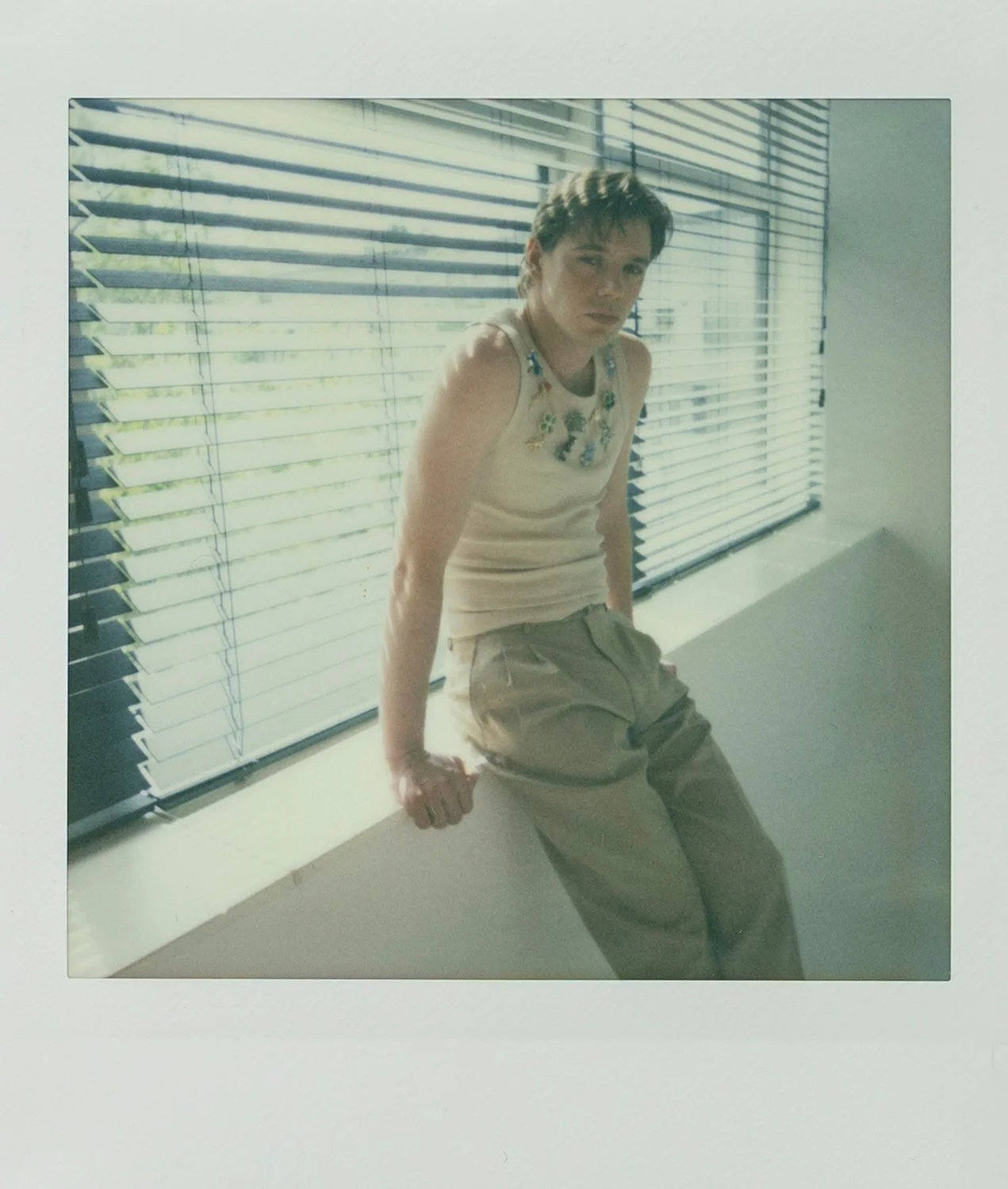 A person sitting on a windowsill in front of horizontal blinds, with light filtering through, wearing a sleeveless shirt with colorful embellishments and beige cargo pants.