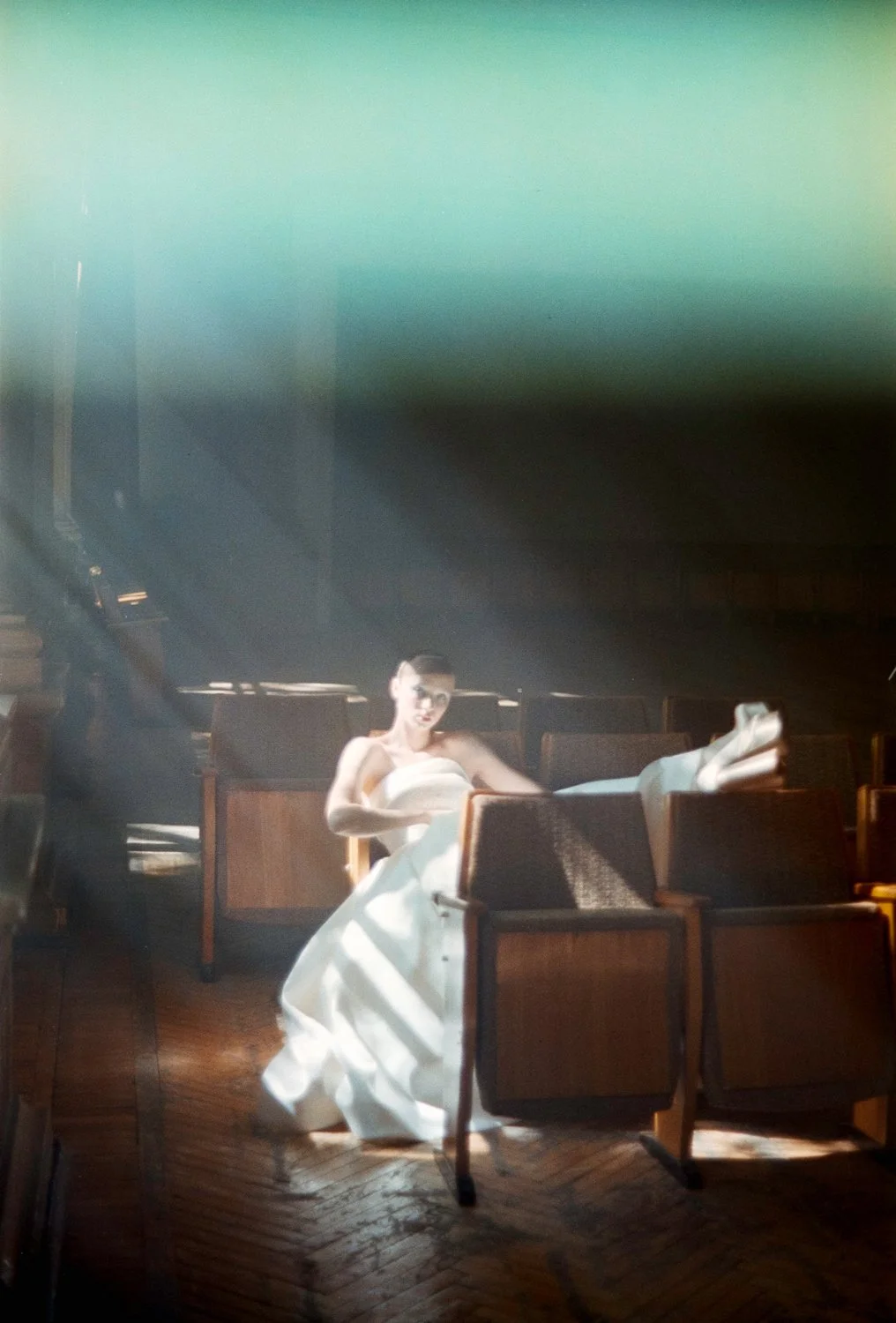 A woman in a white dress sitting alone in a dimly lit room with wooden floors and chairs, illuminated by a beam of light.
