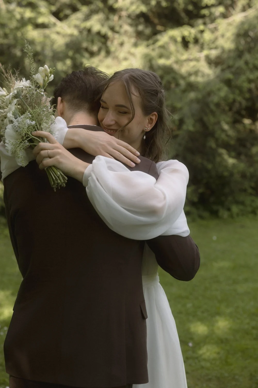 A woman in a white dress hugs a man in a dark suit, holding a bouquet of white flowers, outdoors with lush greenery in the background.