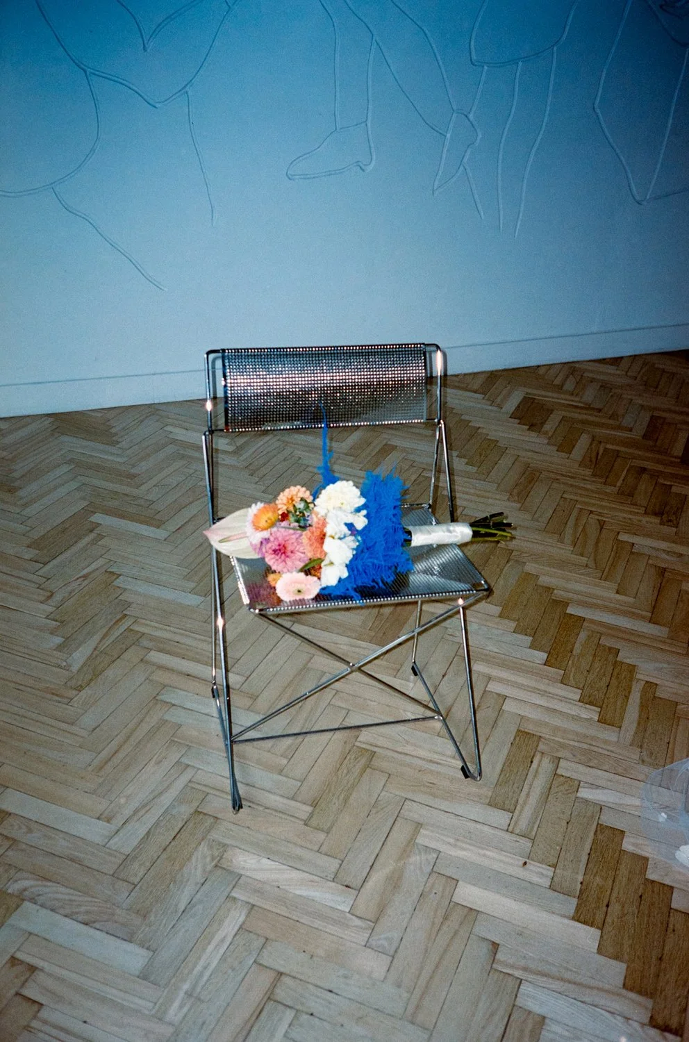 A metal chair with a bouquet of flowers and a rolled-up paper or scroll placed on it, situated on a wooden floor against a painted wall with line drawings.