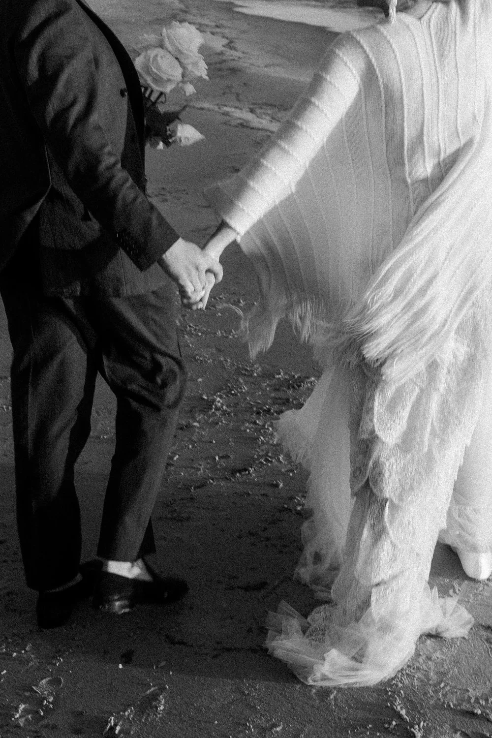 Black and white photo of a couple holding hands during their wedding, with the bride wearing a long, flowing dress and the groom in a suit, on a sandy beach.