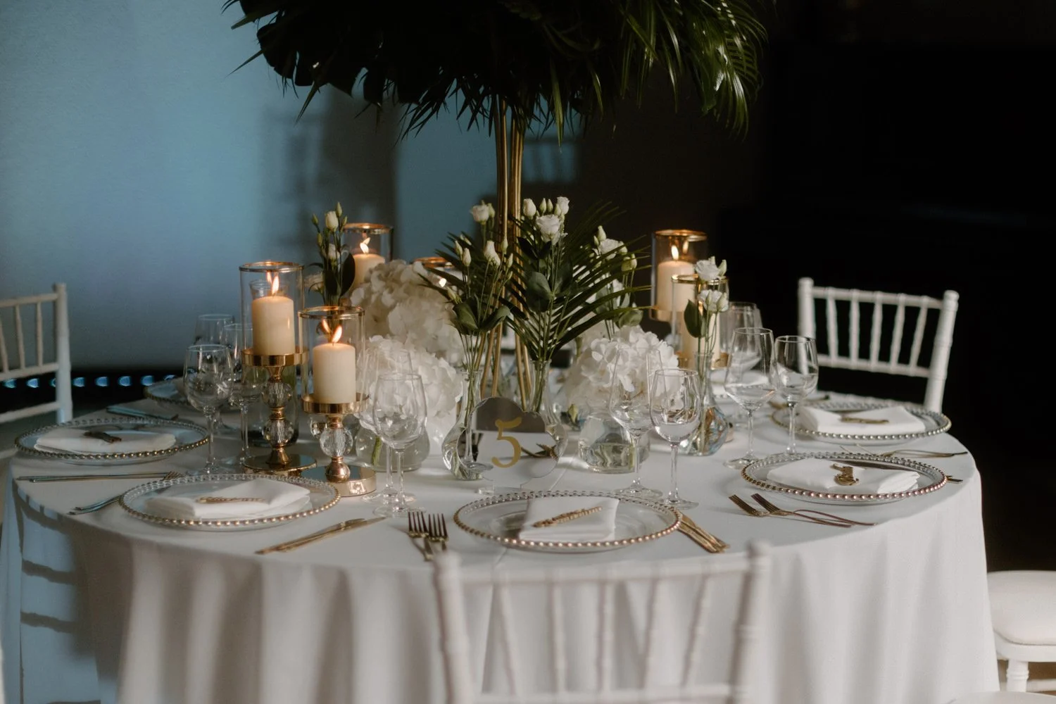 Elegant round table set for a formal event with white tablecloth, gold-rimmed plates, glassware, white napkins, candles, and floral centerpieces, with white chairs around it.