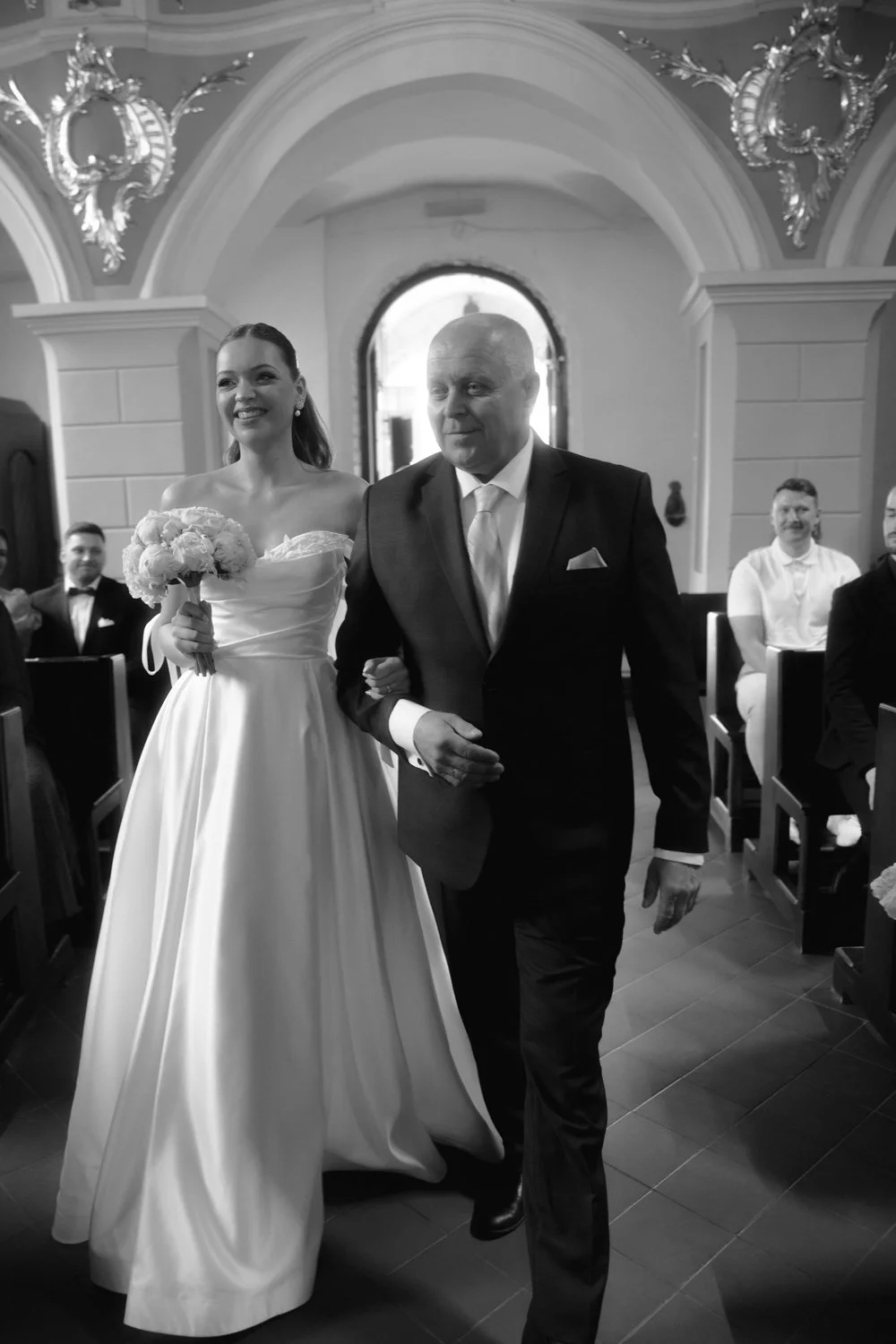 A bride in a white wedding gown holding a bouquet, walking down the aisle with an older man in a dark suit, possibly her father, during a wedding ceremony inside a church.