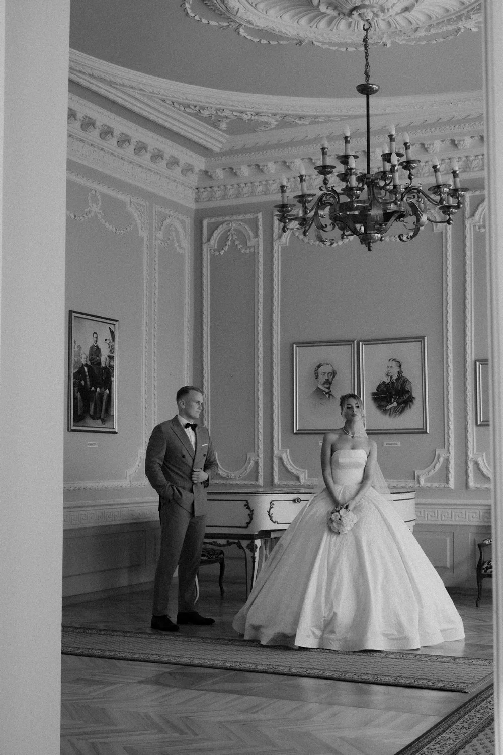 A bride in a strapless wedding gown holding a bouquet, and a groom in a suit with a bow tie, standing in an elegant, ornate room with framed portraits, a chandelier, and decorative wall molding.