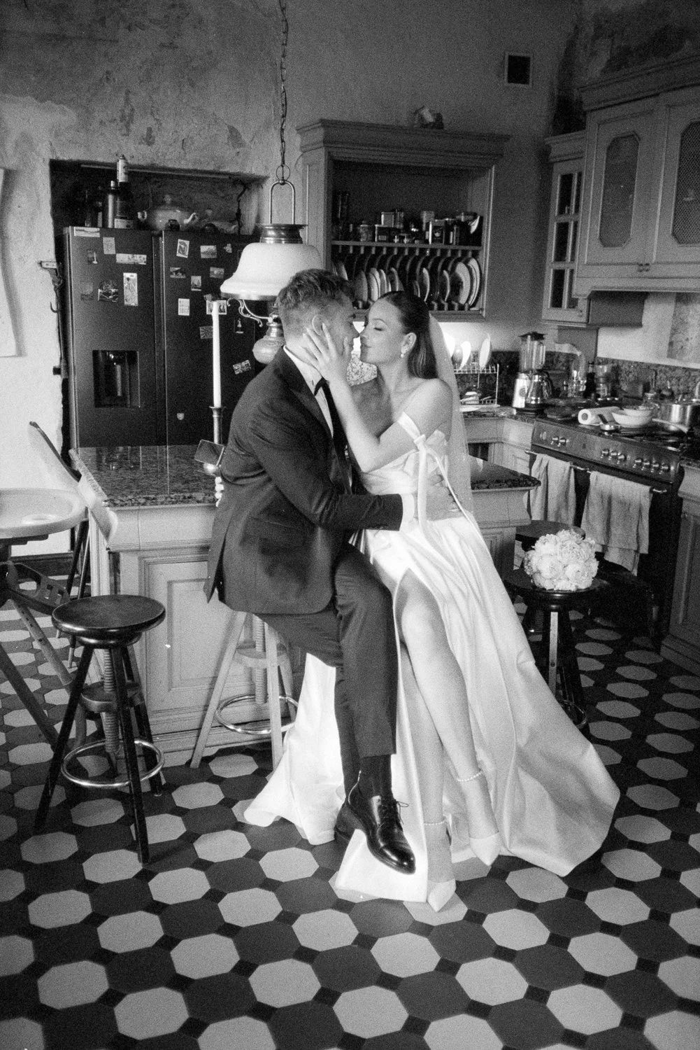 A black and white photo of a bride and groom sharing an intimate moment in a rustic kitchen. The bride is wearing a wedding dress and veil, sitting on a high stool with her legs crossed, while the groom in a suit is sitting on a stool, holding her fa