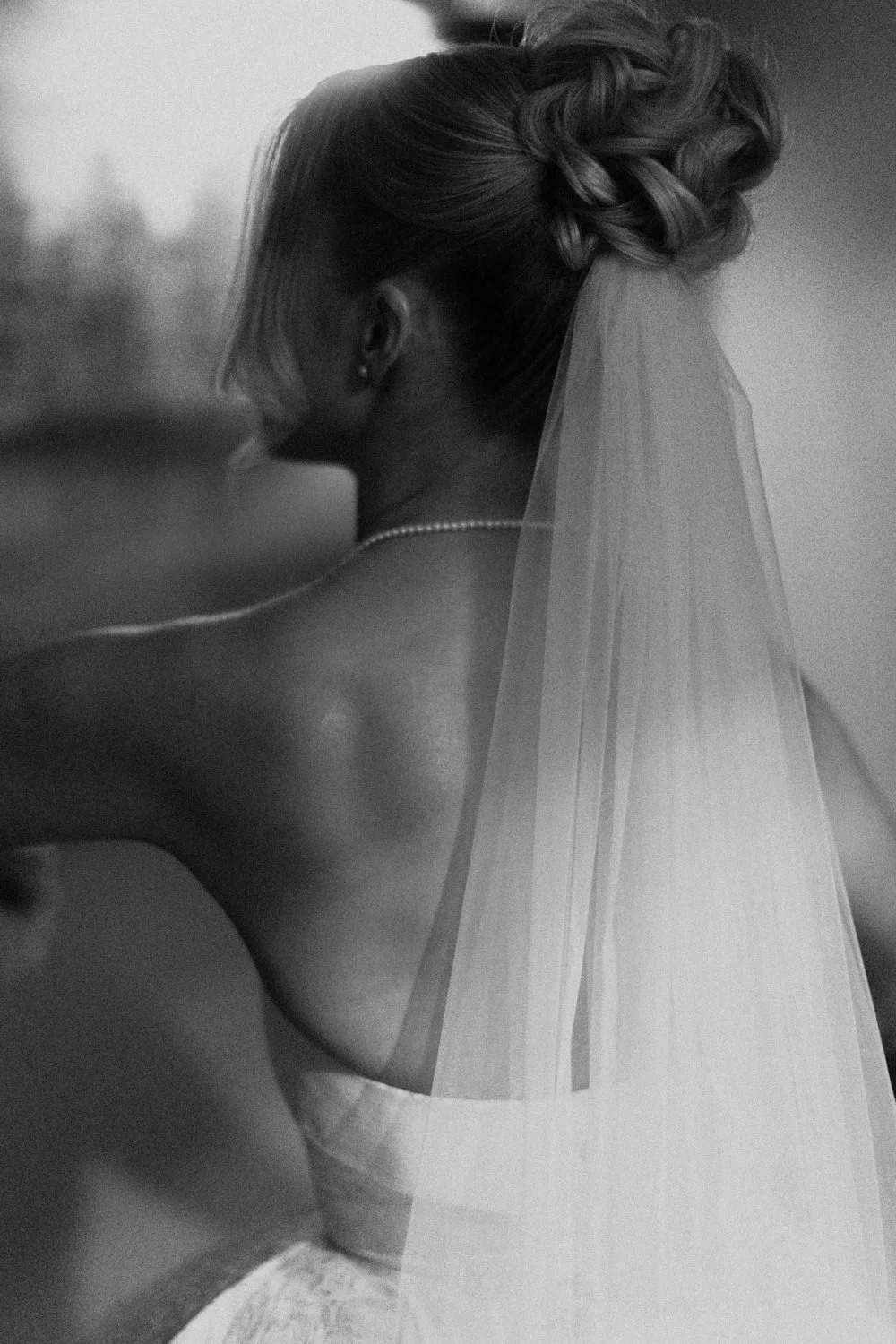 Black and white photo of a bride viewed from behind, with her face turned away. She has braided hair, a pearl necklace, and a sheer veil flowing down her back.