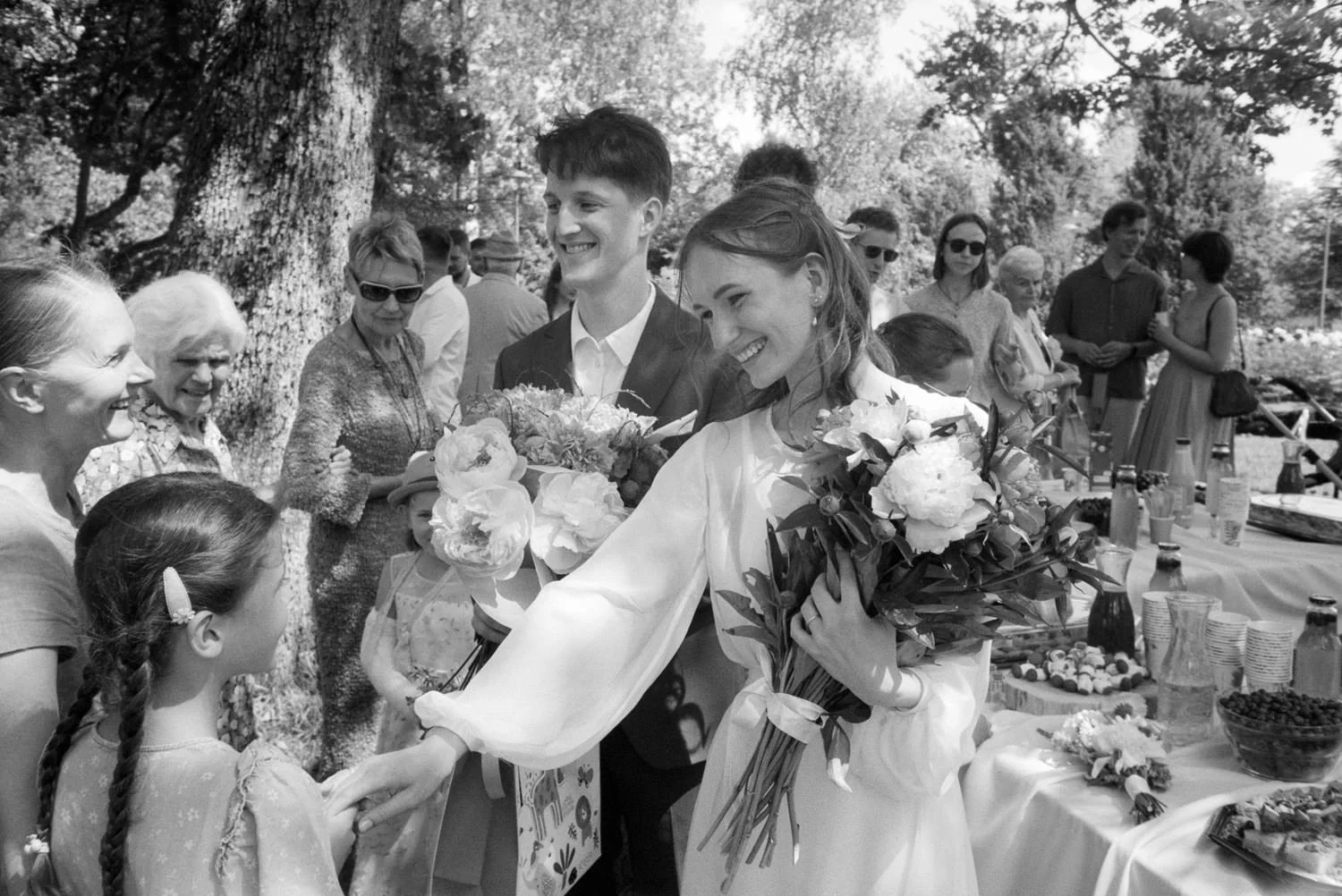 A black and white photo of a wedding celebration outdoors, featuring a bride with long hair and a wedding dress, smiling and handing something to a young girl with pigtails, surrounded by family and friends under trees.