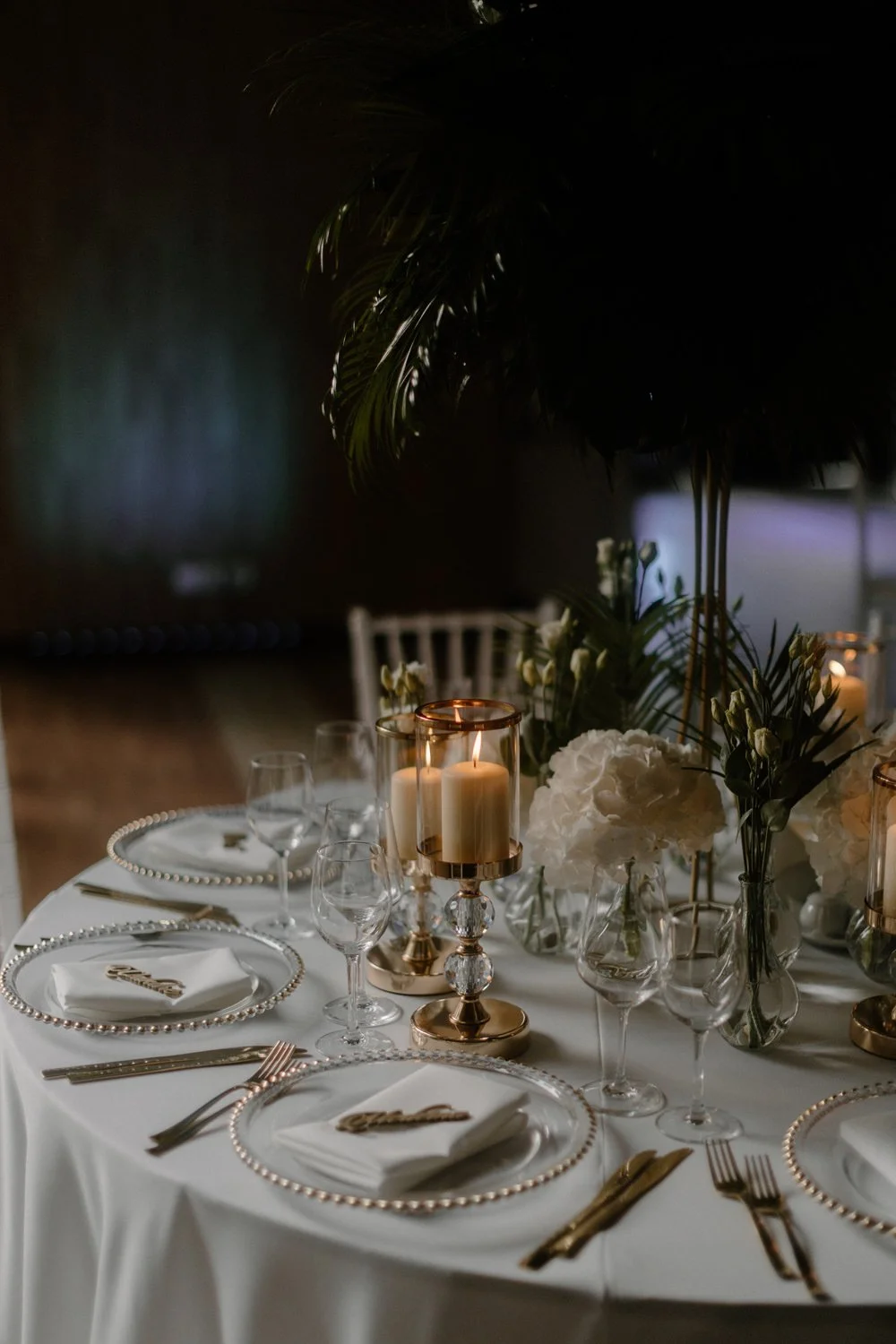 A formal dining table decorated with white flowers, candles, and glassware, set for a special occasion in a dimly lit room.