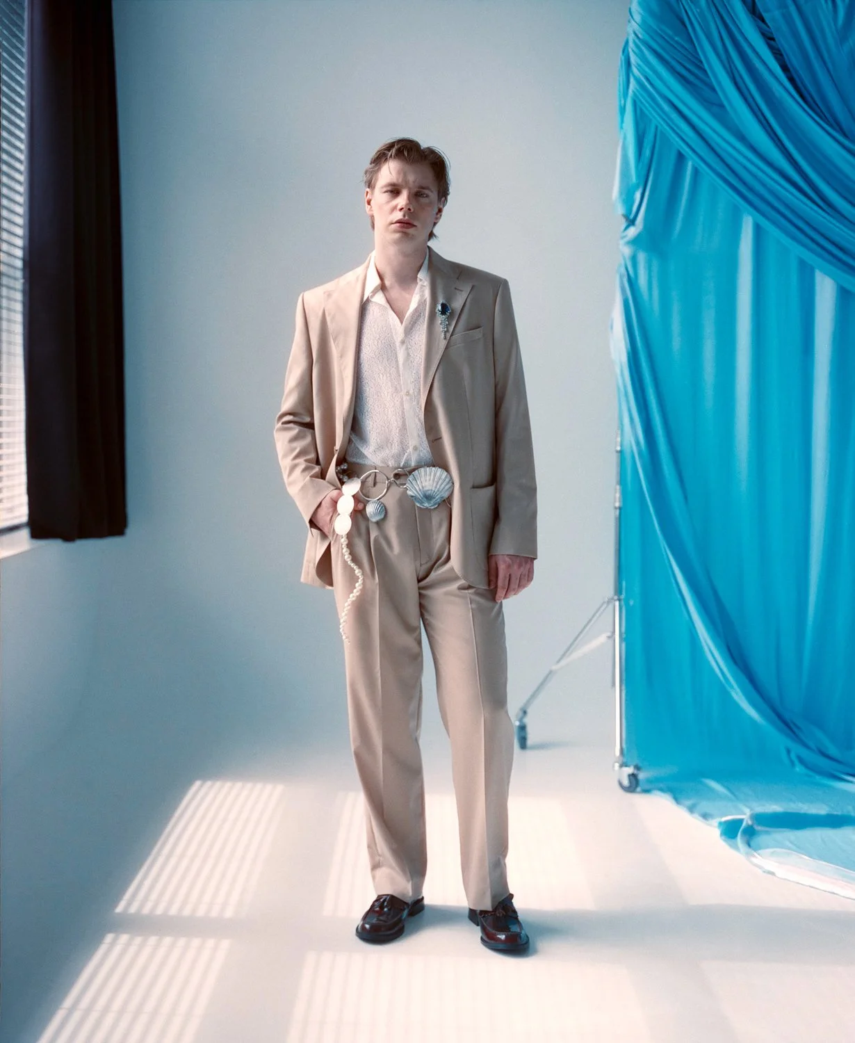 A young man in a beige suit standing in a minimalist room with a blue backdrop. He is wearing accessories including a shell-shaped belt, shell earrings, and a brooch, with a string of beads hanging from his pocket. His expression is neutral, and he i