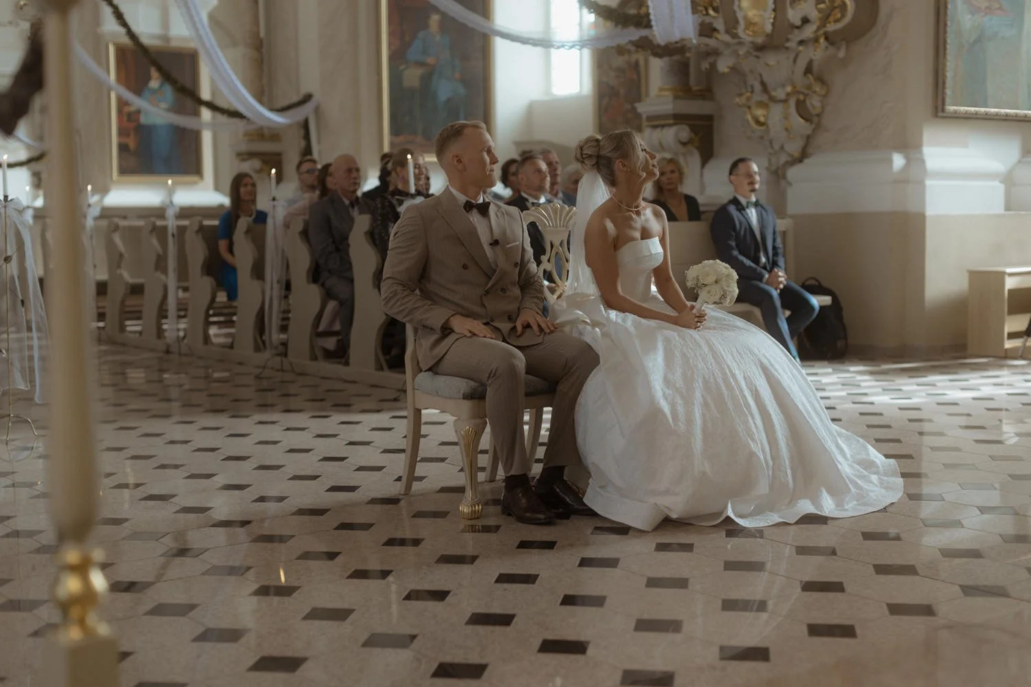 A bride and groom sitting during a wedding ceremony in a church, with guests seated behind them, ornate decorations, and paintings on the walls.