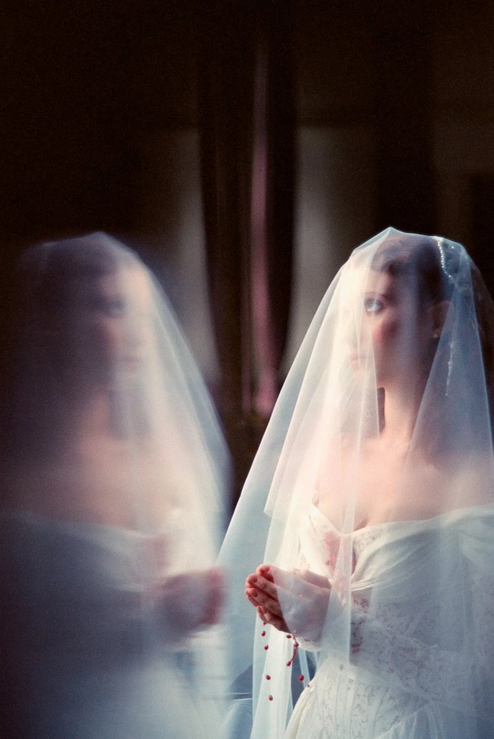 A bride in a white lace wedding dress and veil looking at her reflection in a mirror, with her hands clasped in front of her, and some red spots on her dress.