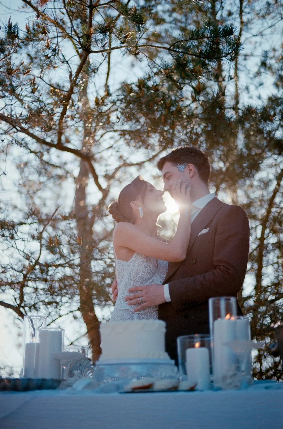 A bride and groom standing outdoors, embracing and touching foreheads, with the sun shining through trees behind them, at a wedding reception with a cake and candles on the table.