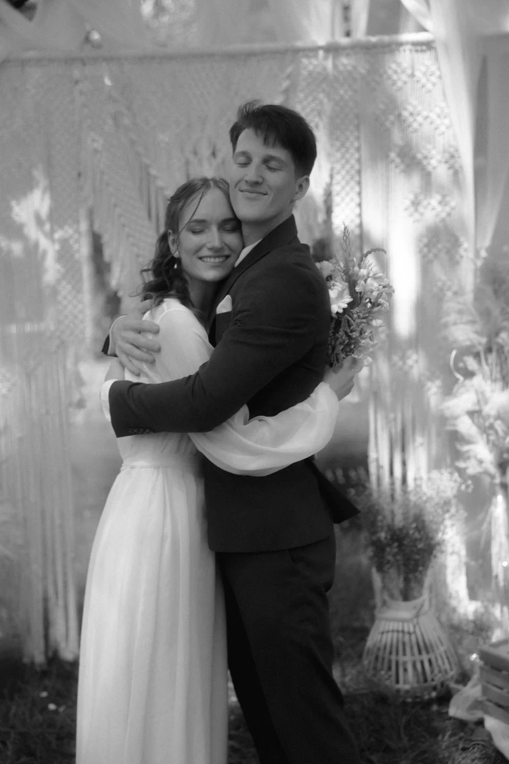 A black and white photograph of a couple hugging at their wedding, with the woman in a white dress and the man in a dark suit, holding a bouquet of flowers.