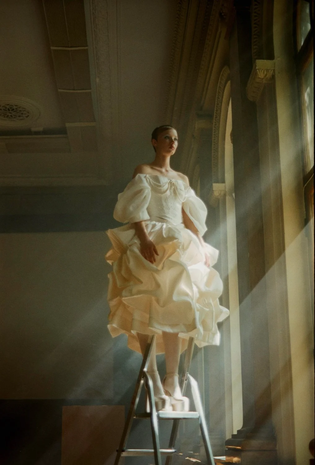 A woman stands on a ladder in a vintage dress, near a large window with sunlight streaming through, with ornate architectural details in the background.