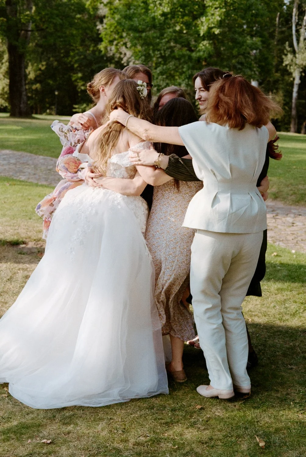 Group of women sharing a hug outdoors, some wearing dresses or suits, in a lush green park with trees in the background.