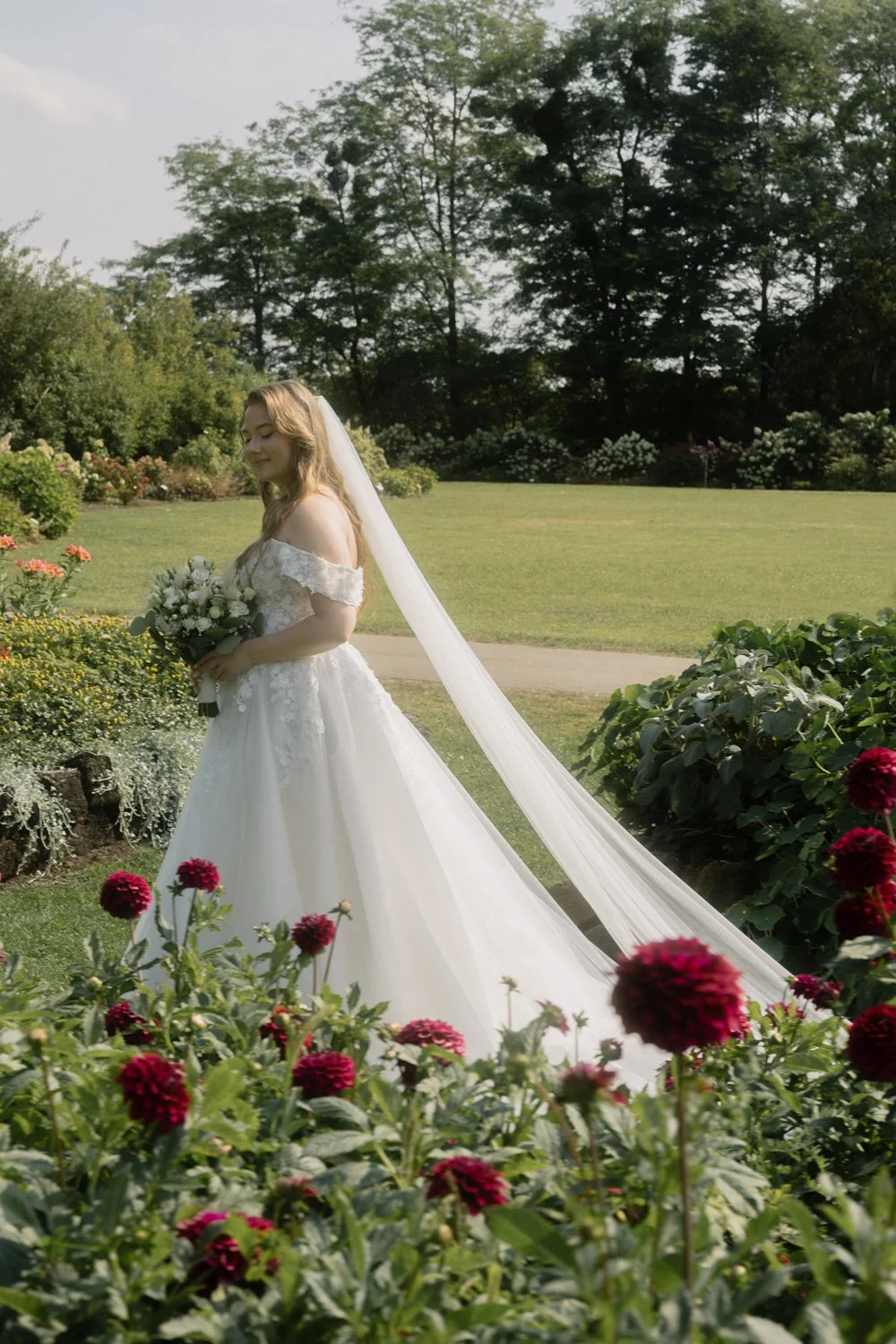 A bride in a white wedding dress holding a bouquet of flowers, standing in a garden with colorful flowers and lush greenery.