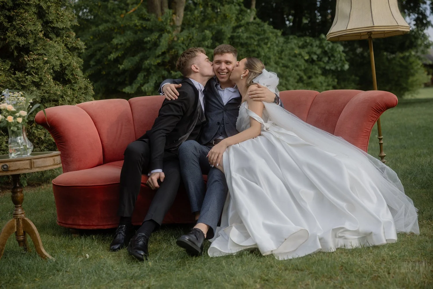 Wedding scene with three people sitting on a red vintage sofa outdoors, surrounded by greenery. The bride, wearing a white wedding dress and veil, is kissing a man in a blue suit, while another man in a black suit is also kissing him on the cheek. Th