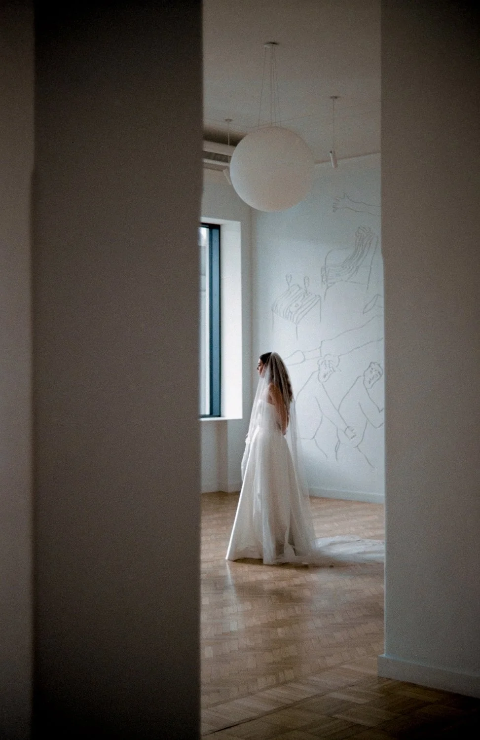 A bride in a white wedding dress and veil standing alone in a softly lit room with modern art on the wall and a large window.