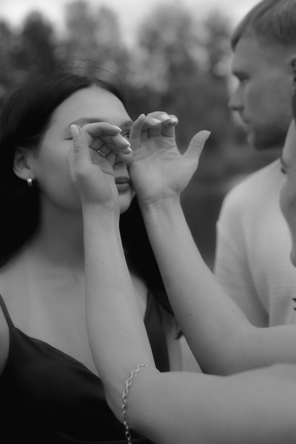 A black and white photo of a woman with long dark hair and earrings, holding her hand near her eye as if looking through a telescope, with a person with short hair and glasses standing next to her, outdoors with blurred trees in the background.