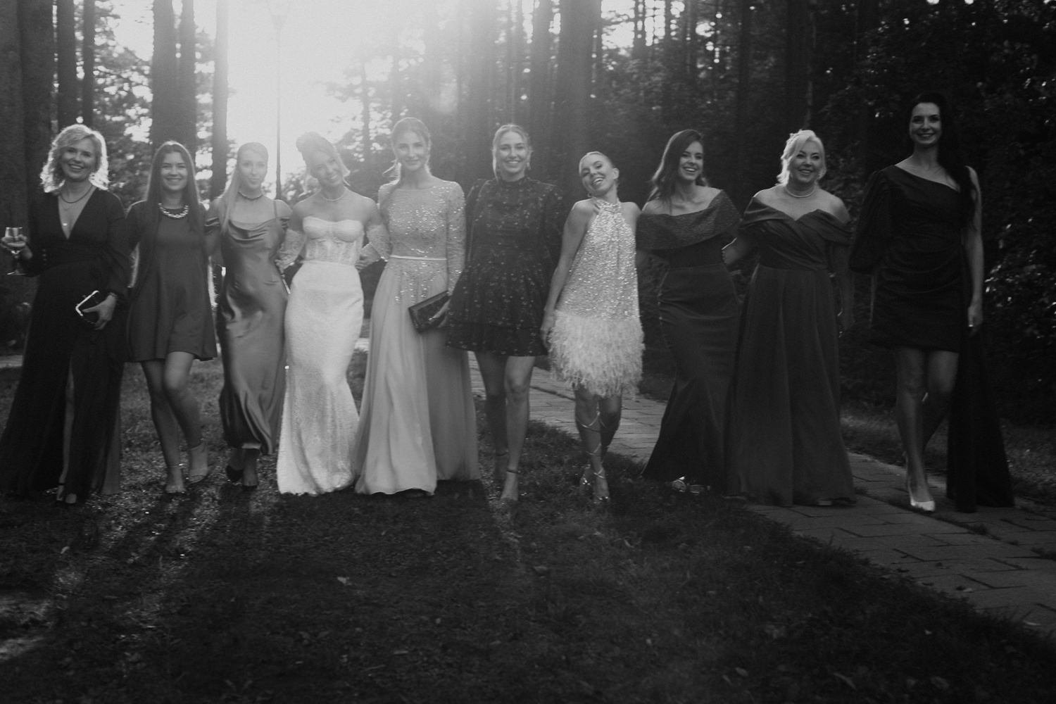 Black and white photo of eleven women in evening gowns standing outdoors on a paved path in a wooded area, smiling and posing for a group photo.