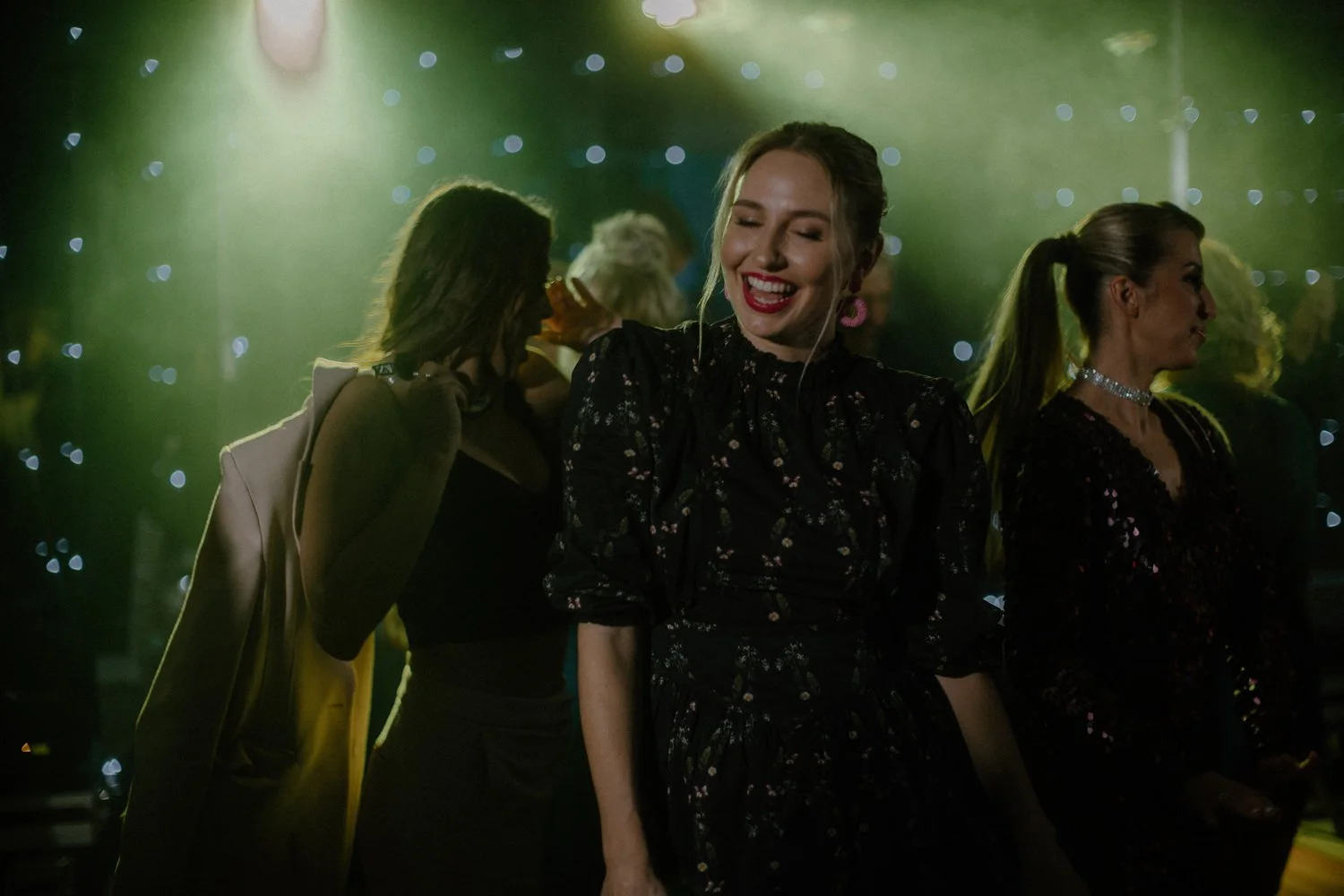 Women dancing and enjoying themselves at a party or club, illuminated by green and yellow lights with bokeh effect in the background.