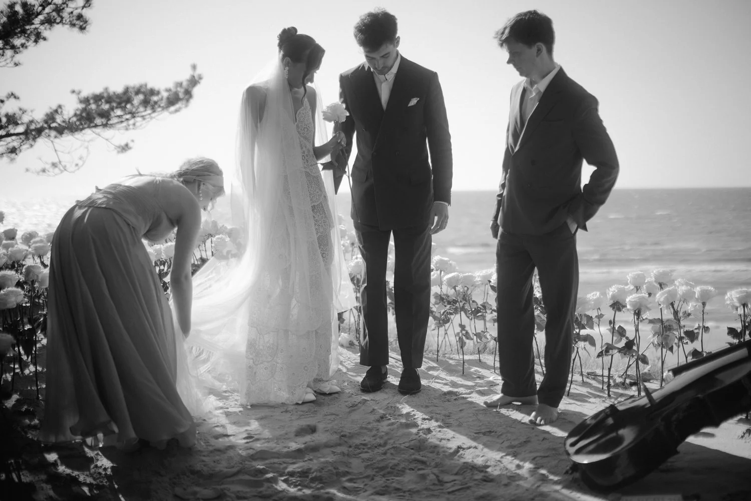 A black and white photo of a wedding scene on a beach with five people: a bride in a white gown holding a flower, a groom in a suit, a bridesmaid adjusting her dress, and another man in a suit standing nearby. The ocean and sky are in the background.