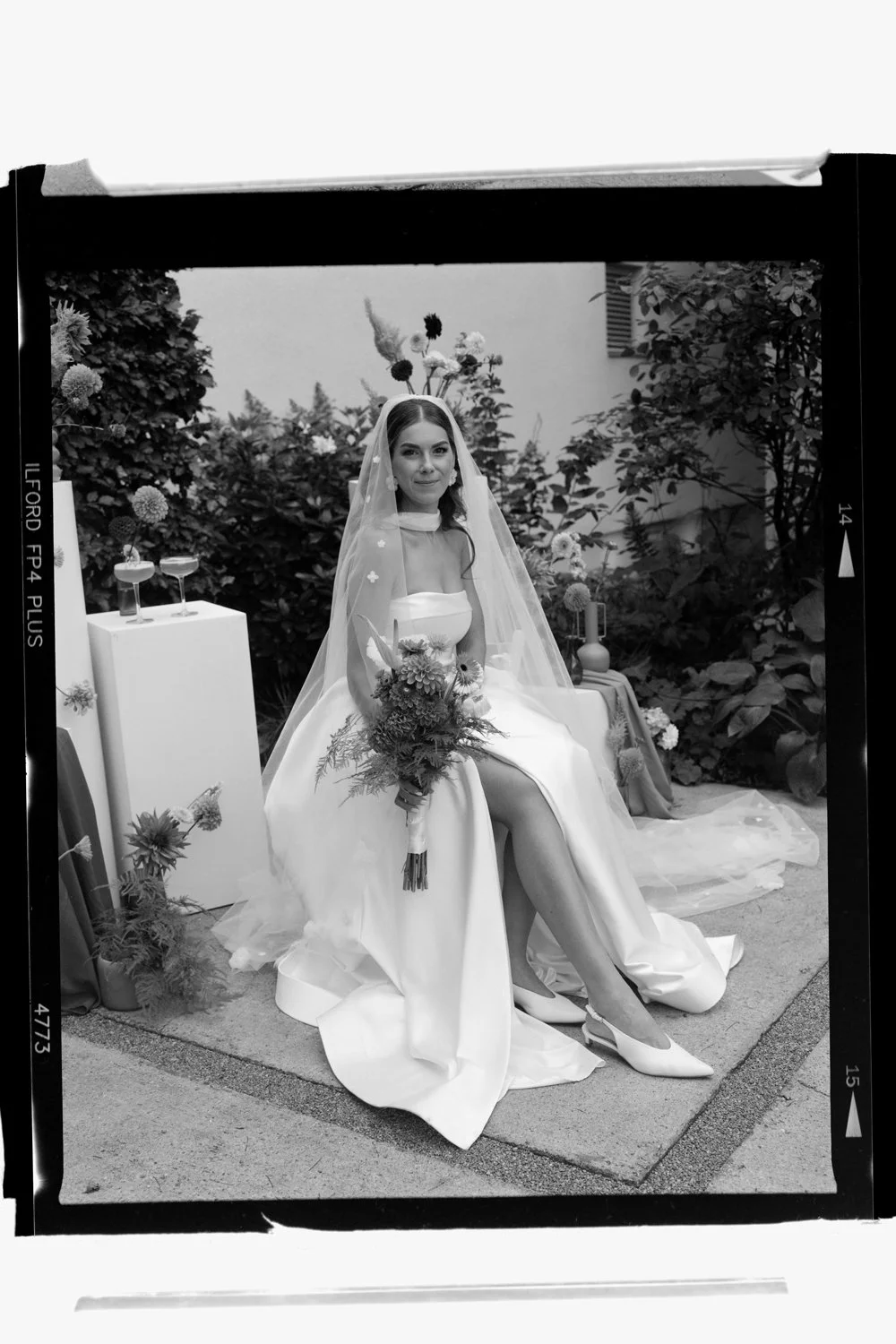 Black and white photo of a bride sitting with her legs crossed, holding a bouquet, wearing a wedding dress and veil, outdoors with flowers and plants around.