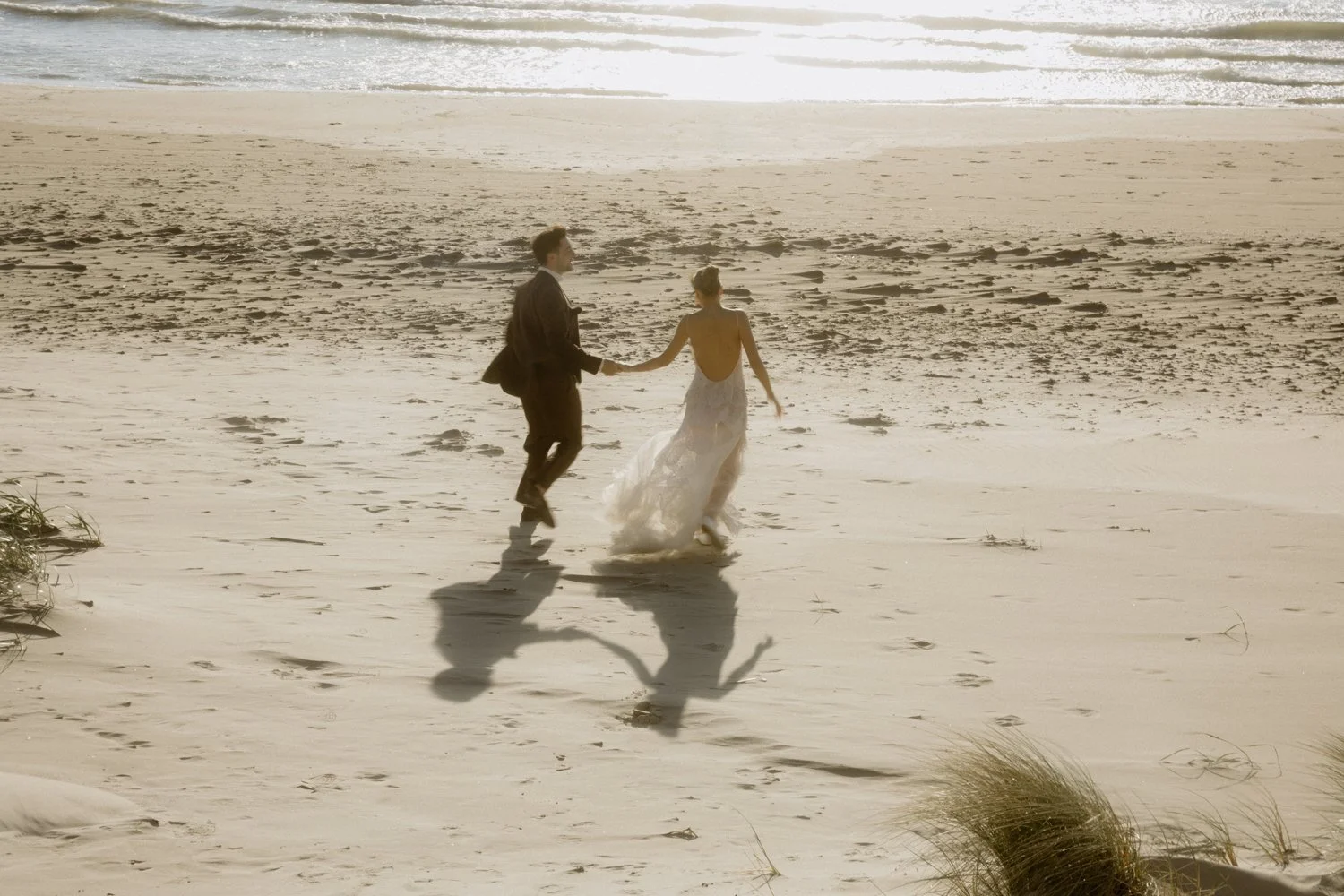 A couple dressed in wedding attire holding hands and running on a beach at sunset, with their shadows visible on the sand.