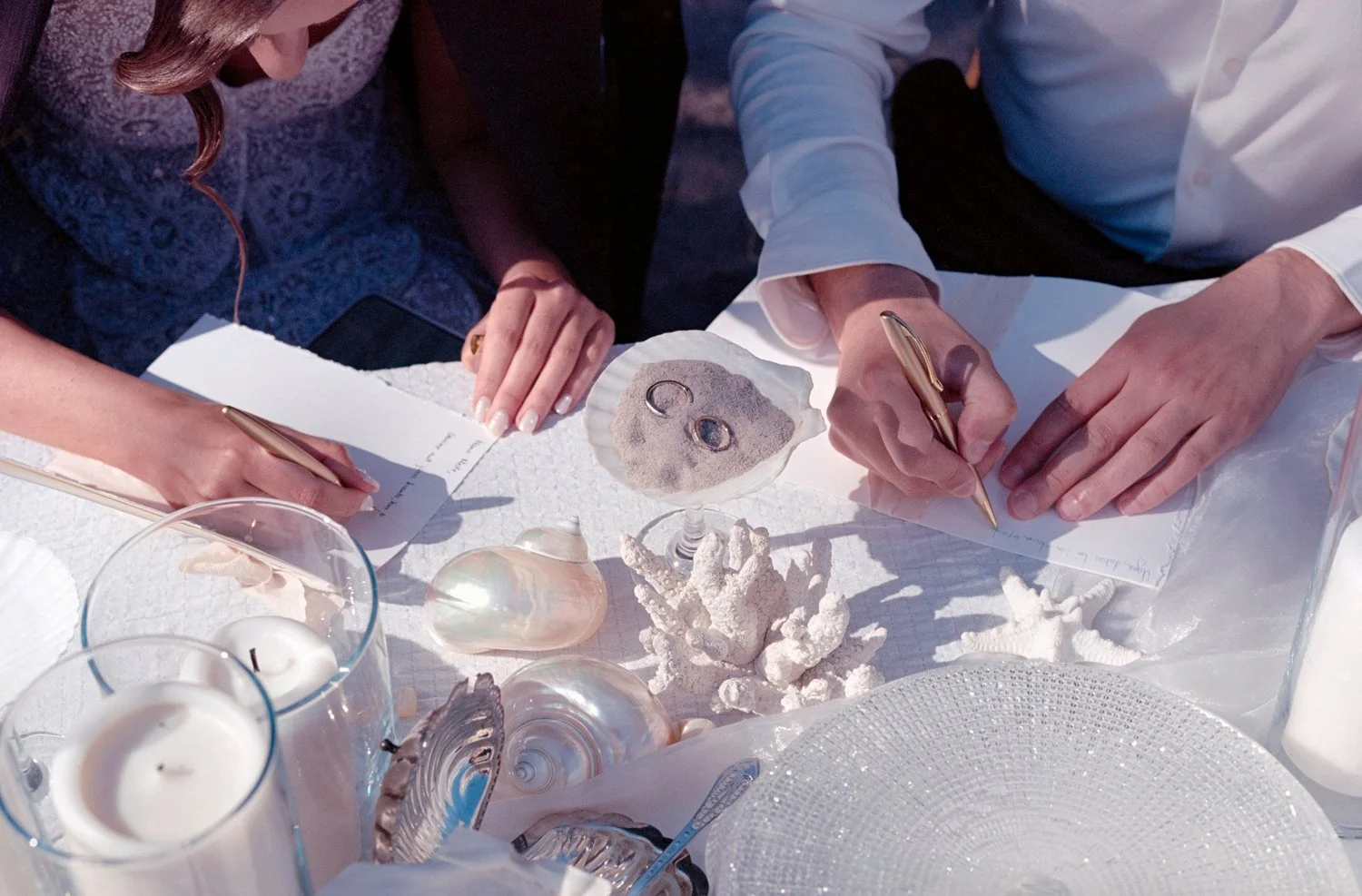 People signing documents on a table decorated with seashells and candles, suggesting a beach or seaside wedding or event.