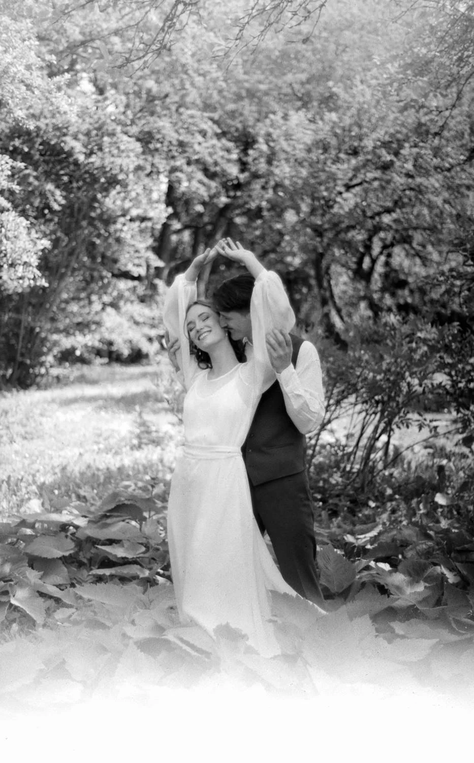 A smiling woman in a white dress and a man in a dark vest and pants dancing outdoors among trees and plants.