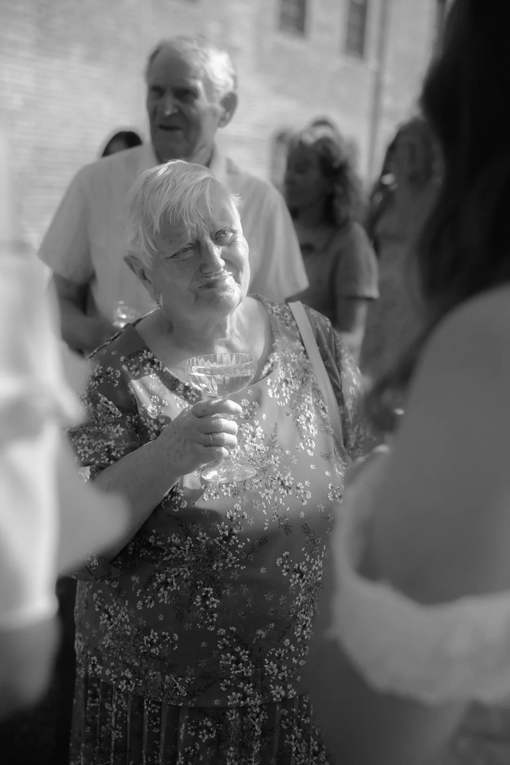 An elderly woman holding a cocktail glass, smiling and engaging in conversation at a gathering. Behind her are other people, some blurry, standing outdoors near a brick wall.