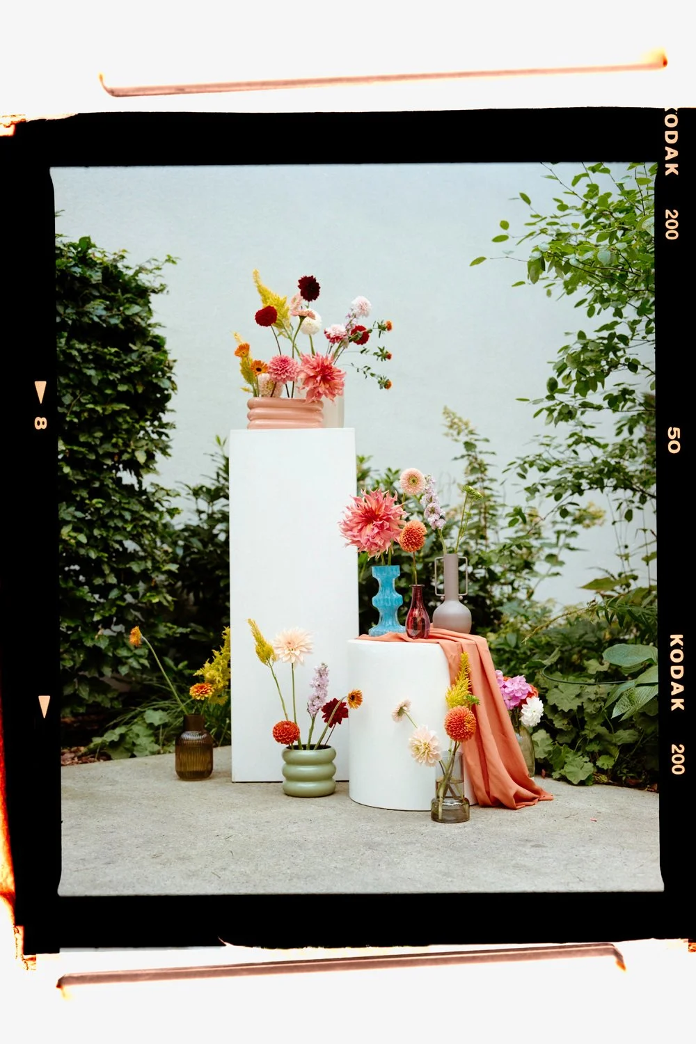 Arrangement of pink, red, yellow, and white flowers in colorful vases on white pedestals and small tables outdoors, surrounded by green foliage.