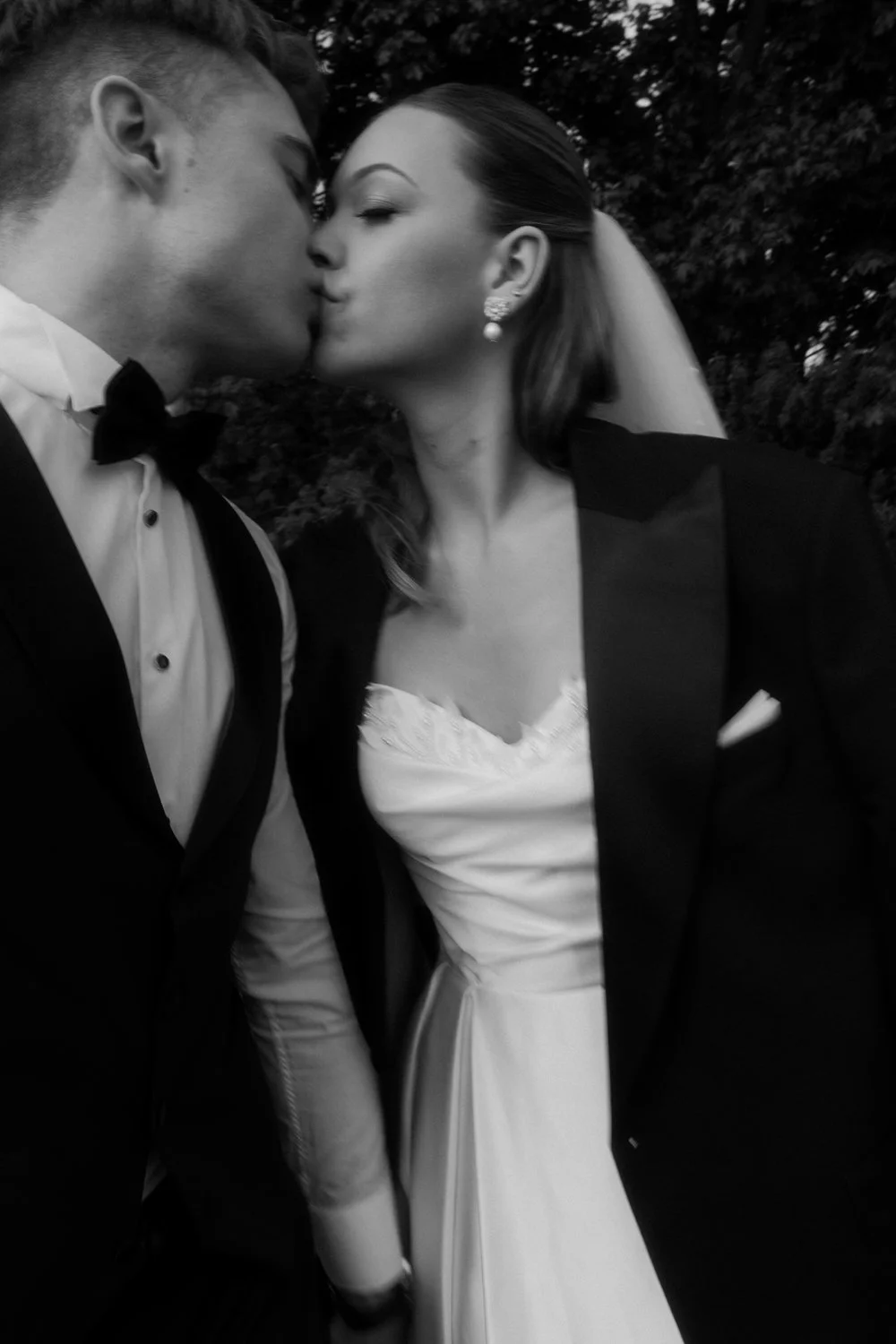 Black and white photo of a couple in wedding attire kissing outdoors, with trees in the background.