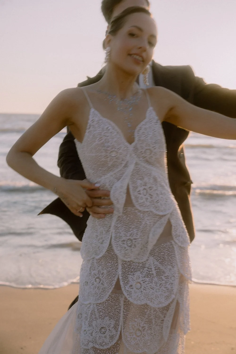 A woman in a white lace wedding dress standing on the beach with a man in a dark suit behind her, holding her waist during sunset.