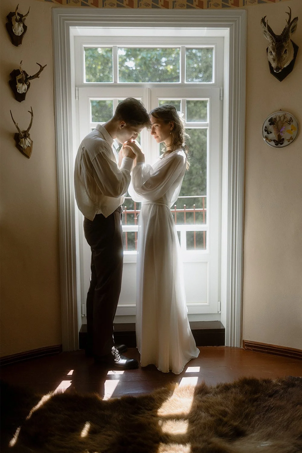 A young weddings couple holding hands and touching foreheads inside a room near a window with sunlight. The woman is wearing a long white dress, and the man is dressed in dark trousers and a light shirt. The room has deer head wall mounts and a butte