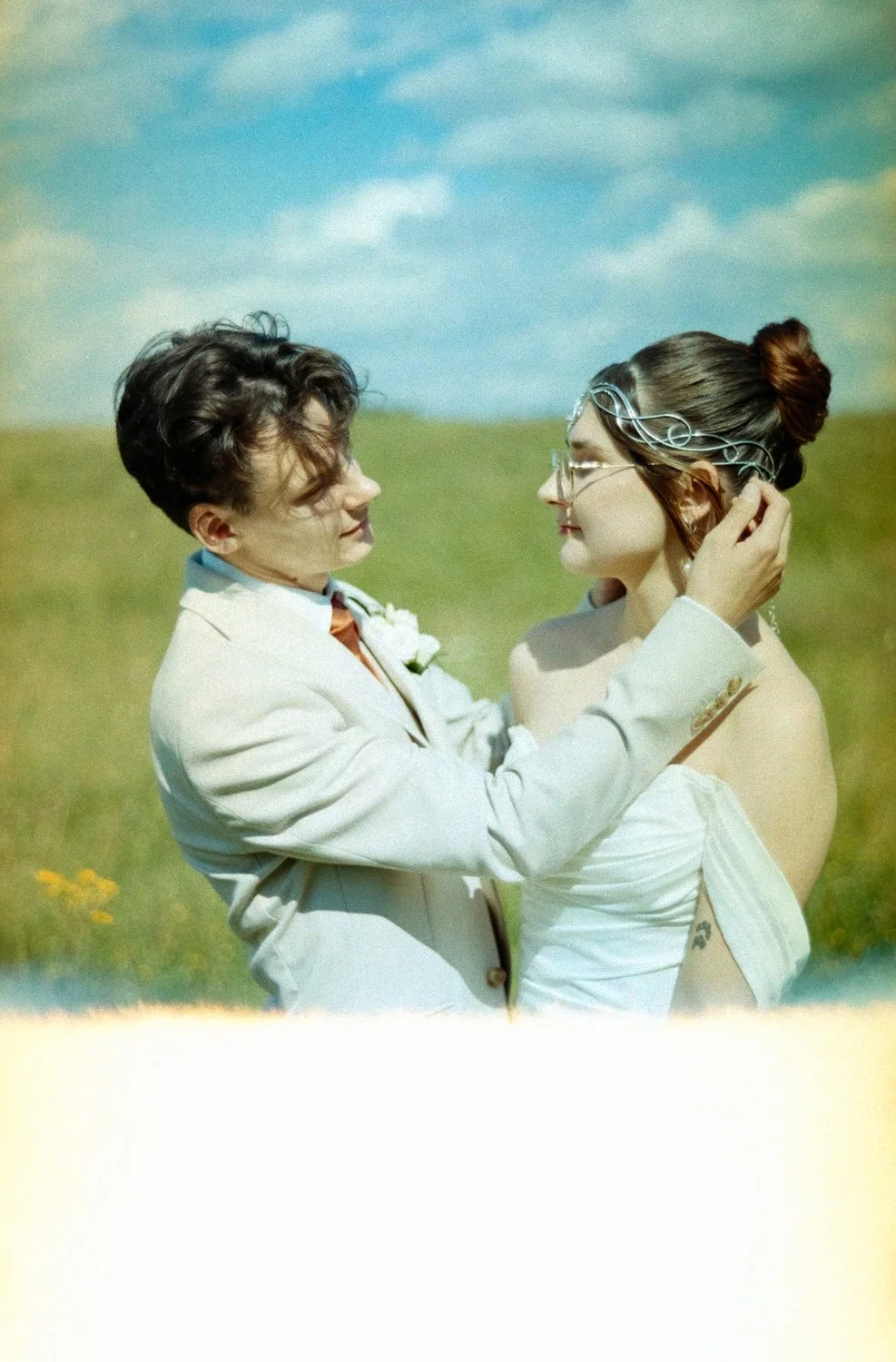 A man and woman in wedding attire standing in a field, gazing at each other. The man is holding the woman's face gently, both smiling. The woman is wearing glasses and has her hair in an updo.