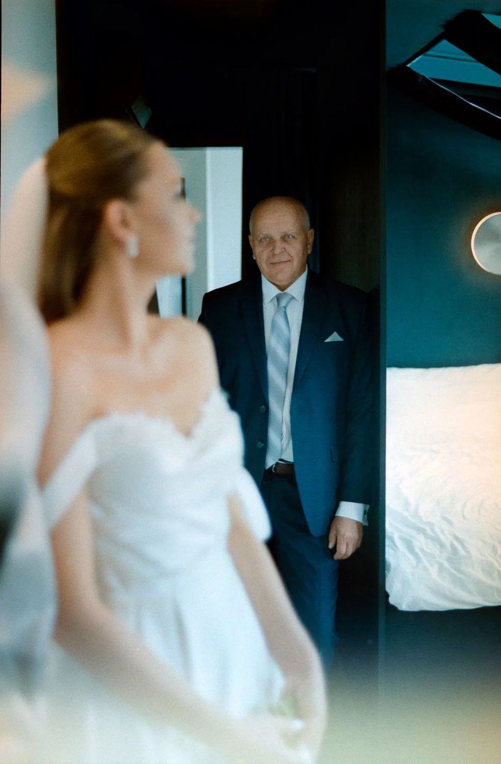 A bride in a white wedding dress and an older man in a dark suit are in a room, with the man looking at the bride.