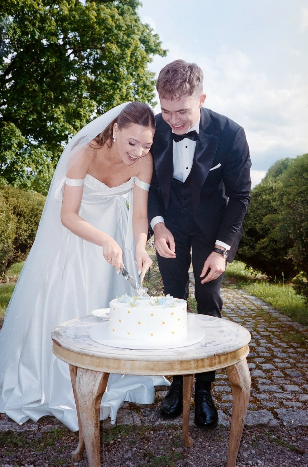 A bride and groom smiling and cutting a wedding cake outdoors on a sunny day.