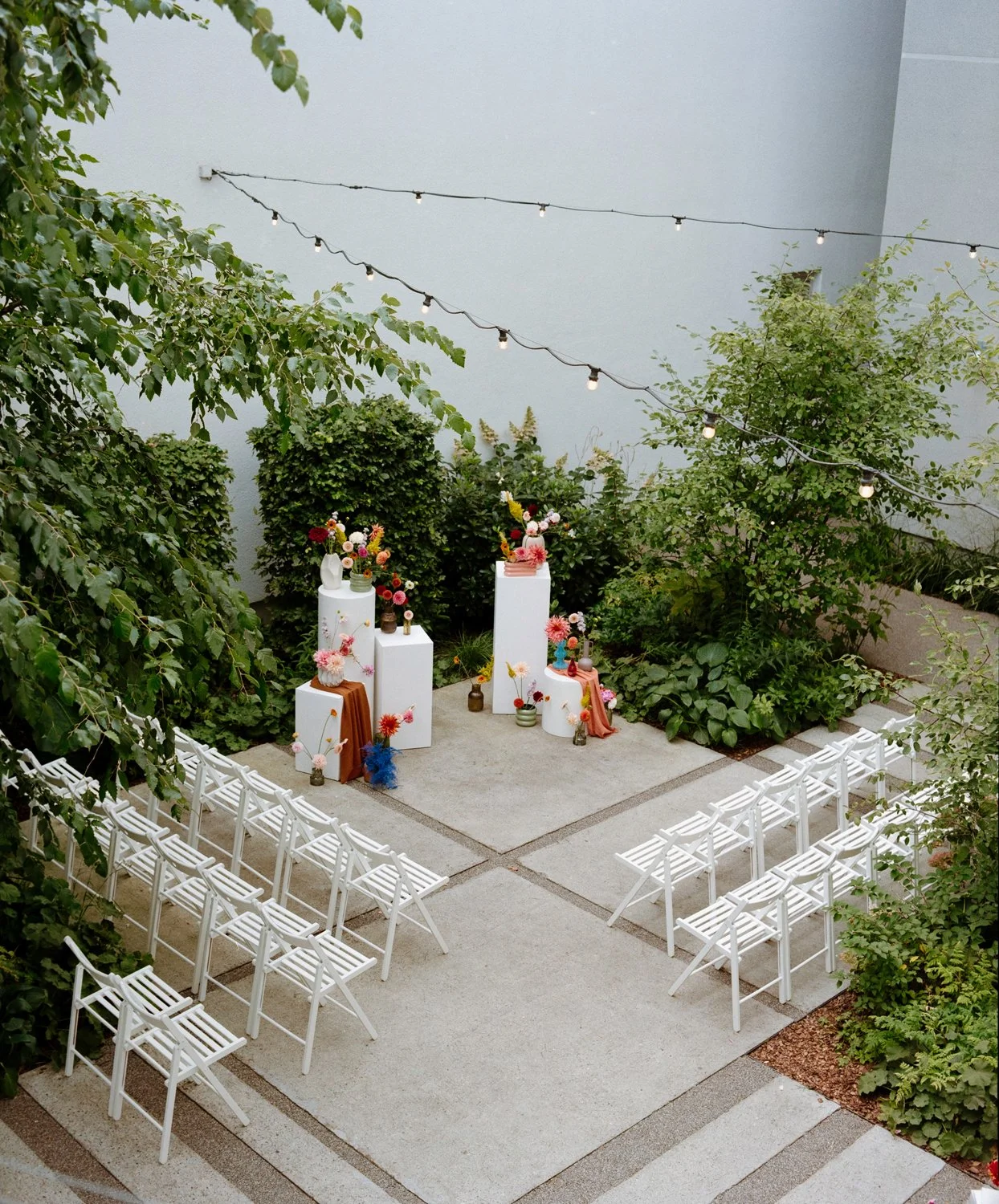 Outdoor wedding ceremony setup with white chairs, floral arrangements, and decorative pedestals in a garden with string lights overhead.
