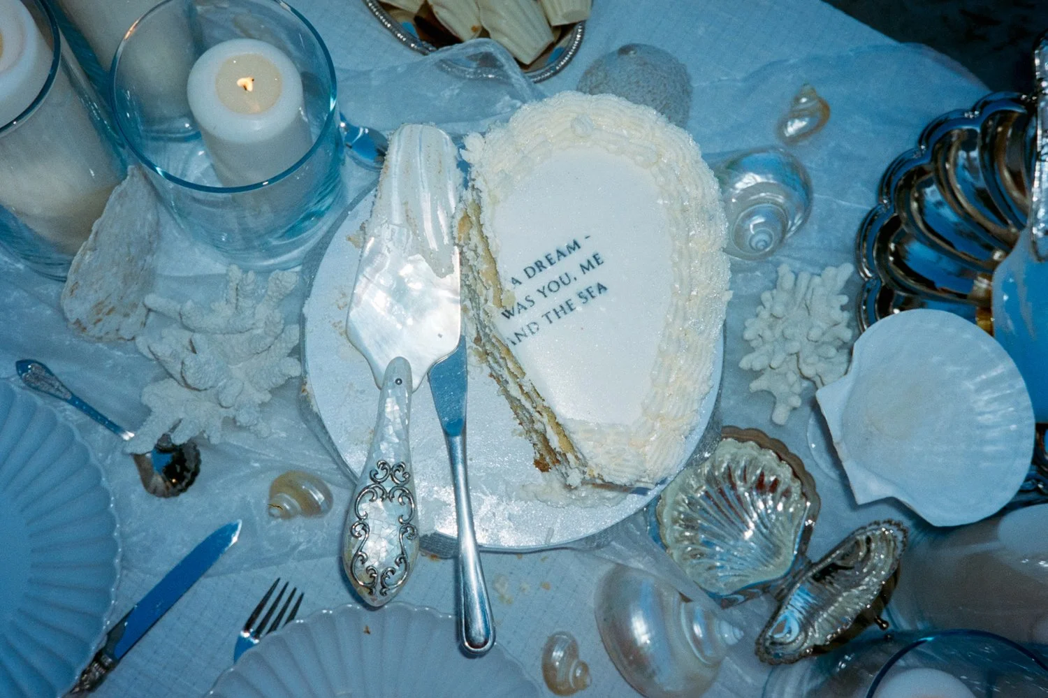 A partially cut white cake with seashell decorations and the message 'A DREAM WAS YOU, ME AND THE SEA' on top, surrounded by seashell-shaped and transparent glass decorations, candles, and silverware on a table with a blue and white theme.