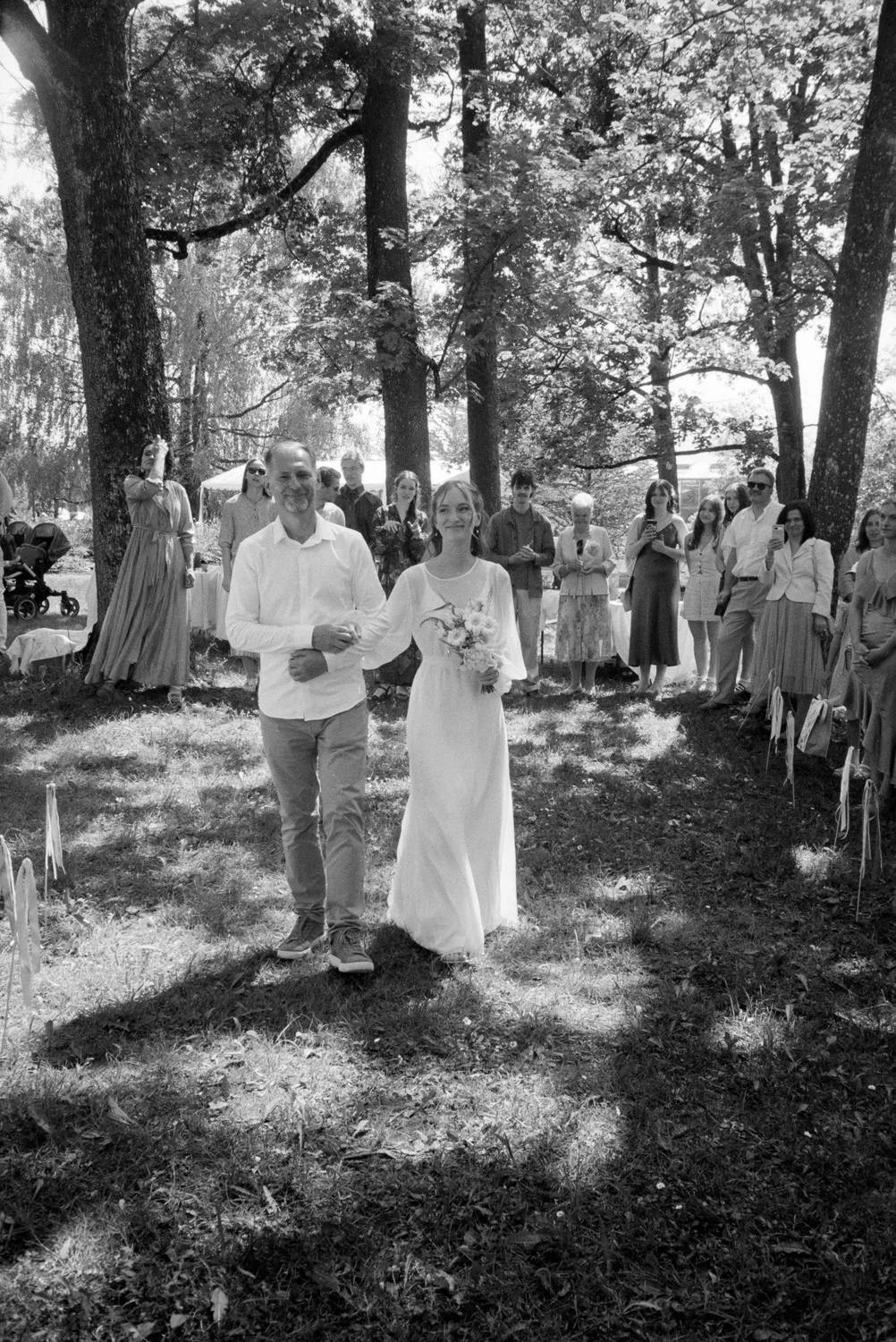 A bride and groom walking down an outdoor aisle surrounded by trees, with guests watching and taking photos.