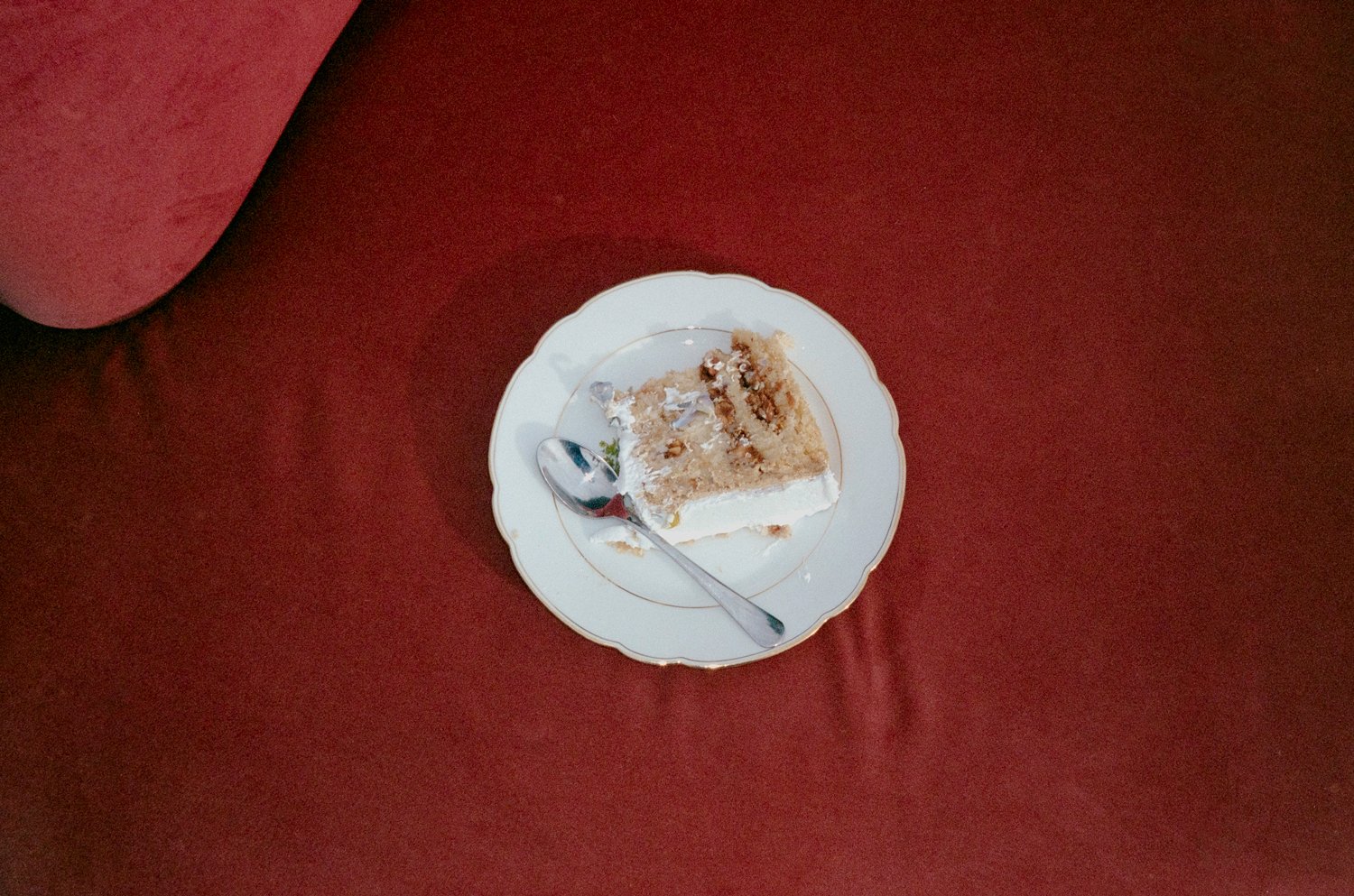 A slice of cake with white frosting on a small decorative plate, with a spoon resting on the plate, placed on a red surface.