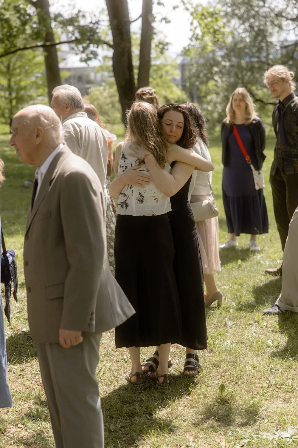 Two young women embracing each other, one with curly hair and the other with straight hair, in a grassy outdoor setting during daytime, surrounded by other people.