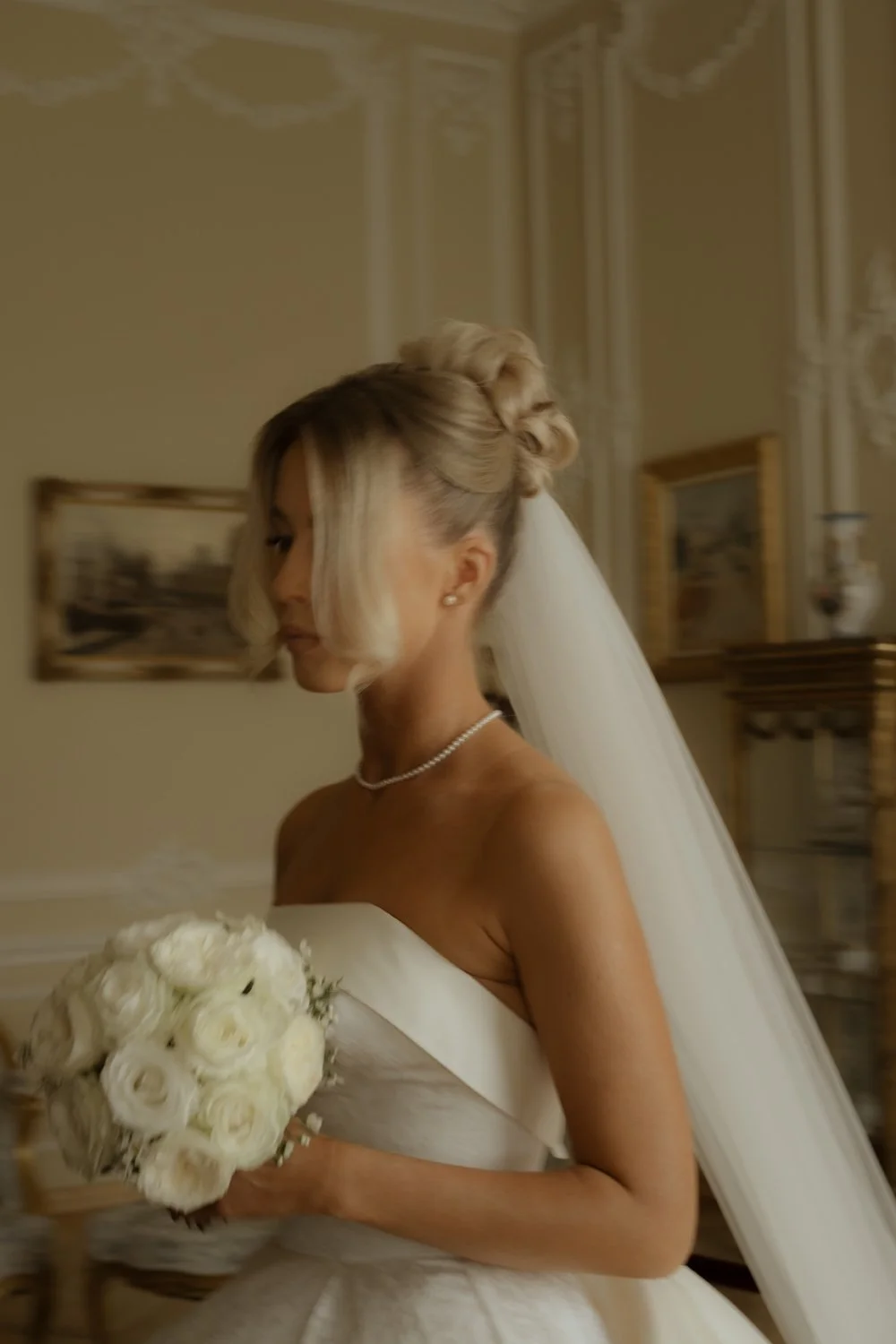 Bride with styled blonde hair, wearing pearl earrings and necklace, holding a white bouquet of roses, dressed in an off-shoulder wedding gown, standing indoors in a decorated room.