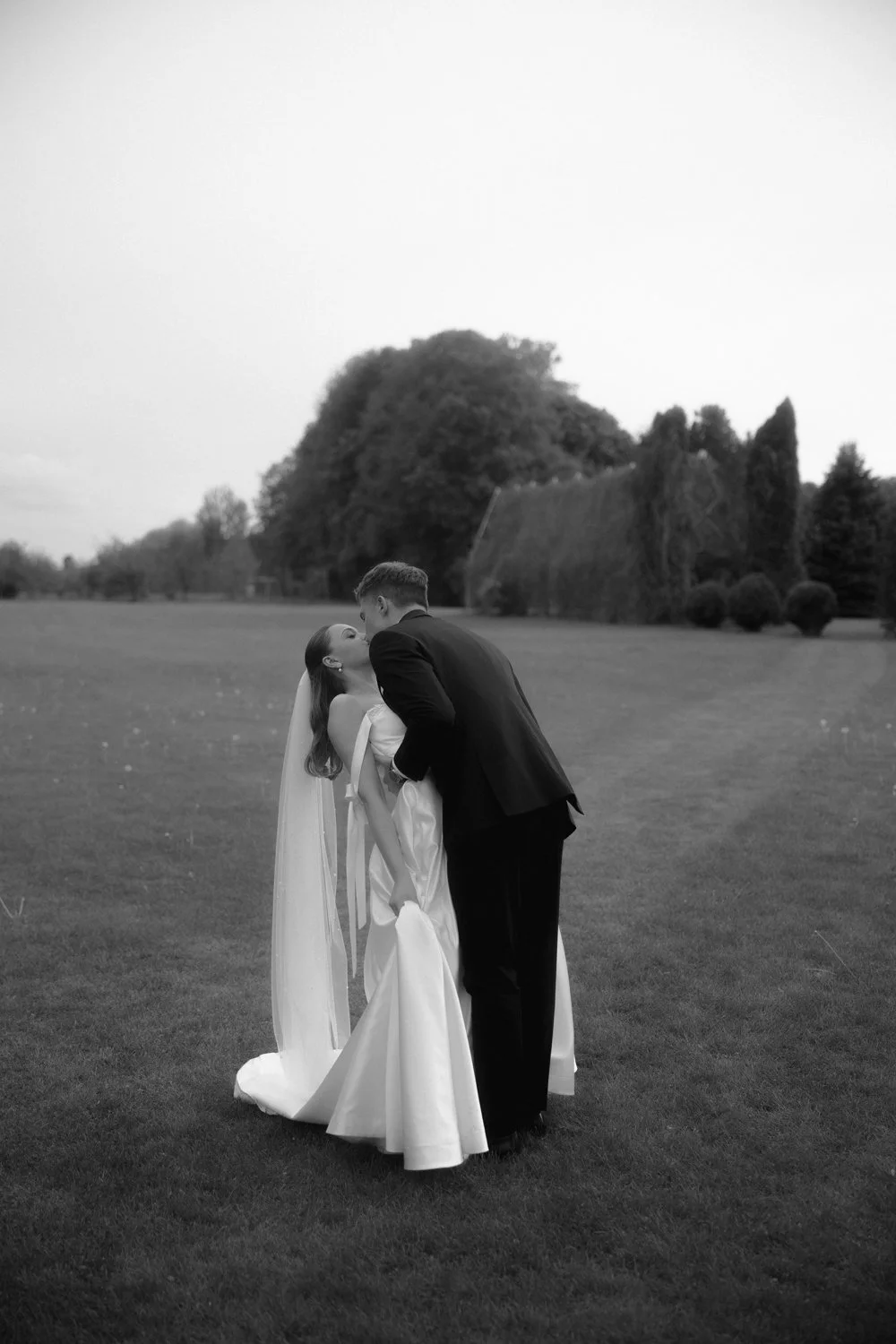 A black-and-white photo of a bride and groom kissing outdoors on a grassy field with trees in the background.