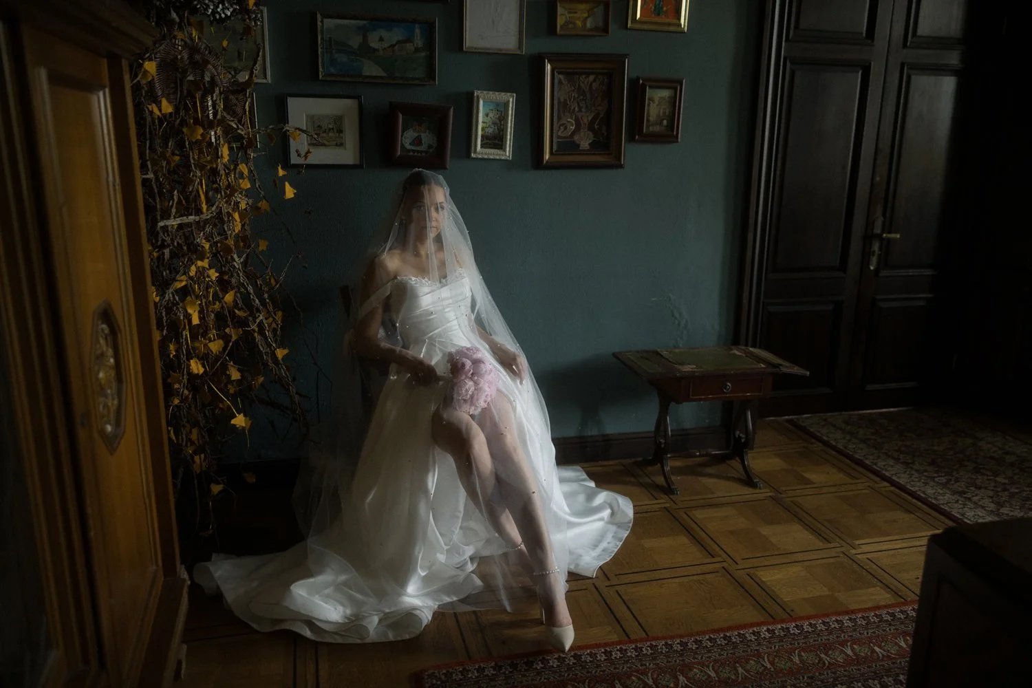 A woman dressed in a wedding gown with a veil, sitting on a chair in a dimly lit room, holding a bouquet of pink flowers, with artwork hanging on the wall behind her.